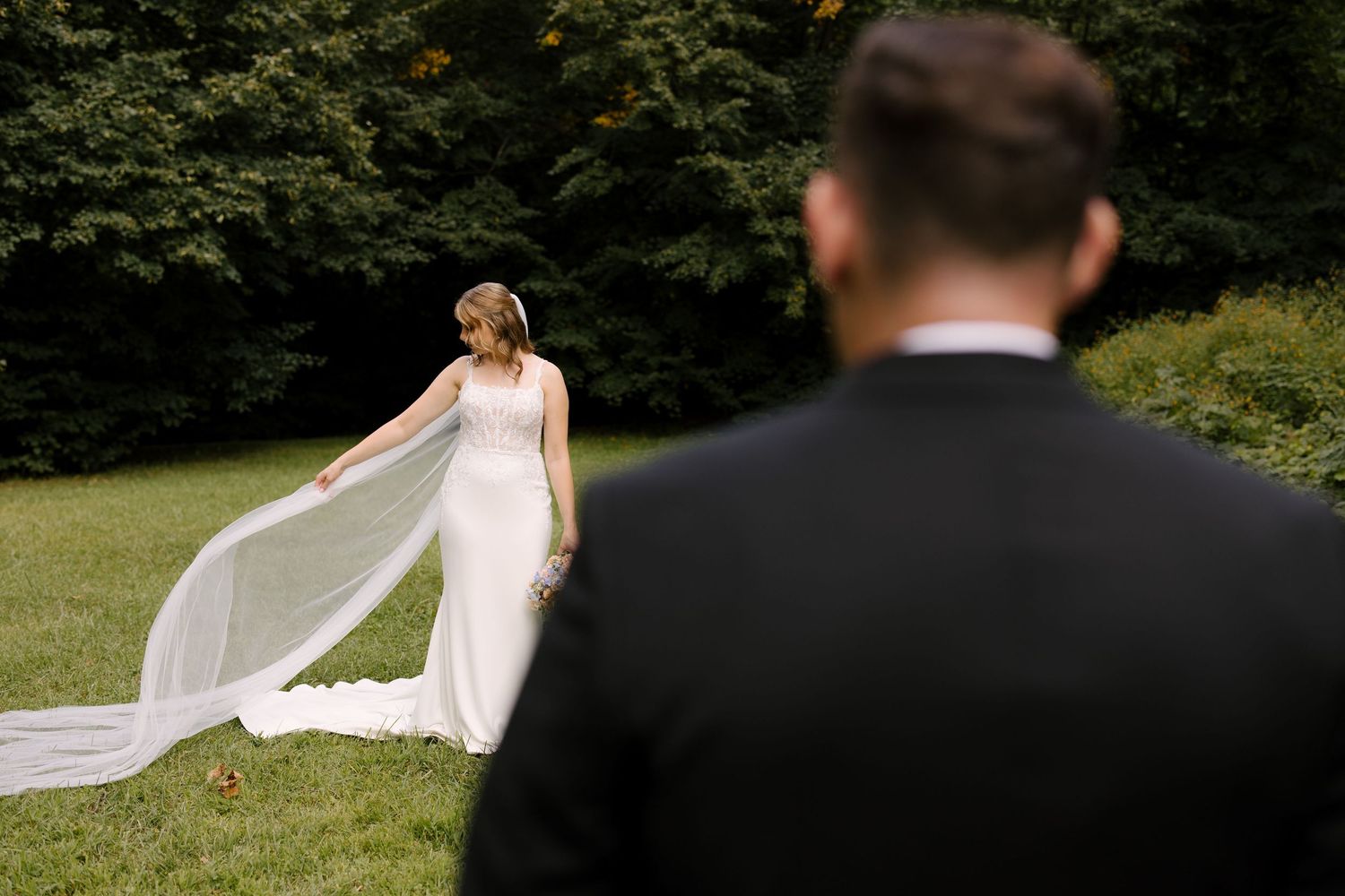 Bride in fitted white gown with flowing veil walks toward groom on lush green lawn.