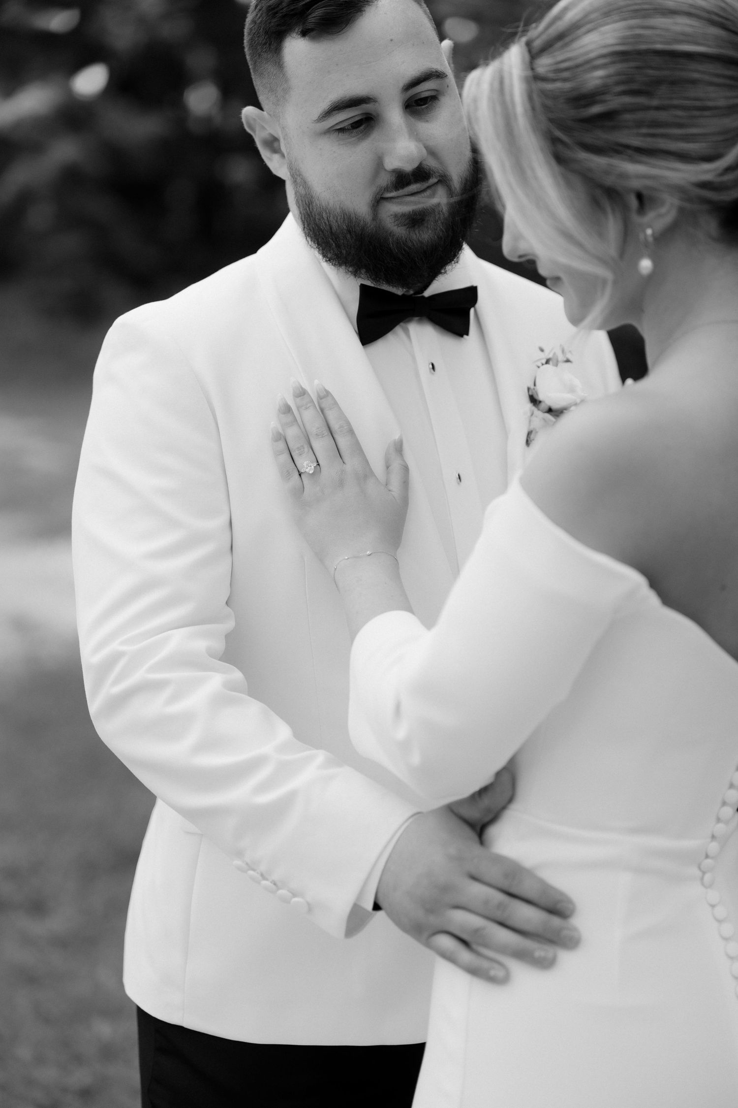 Emotional groom in white tuxedo reacting with tears as bride places hand on his chest during first look.