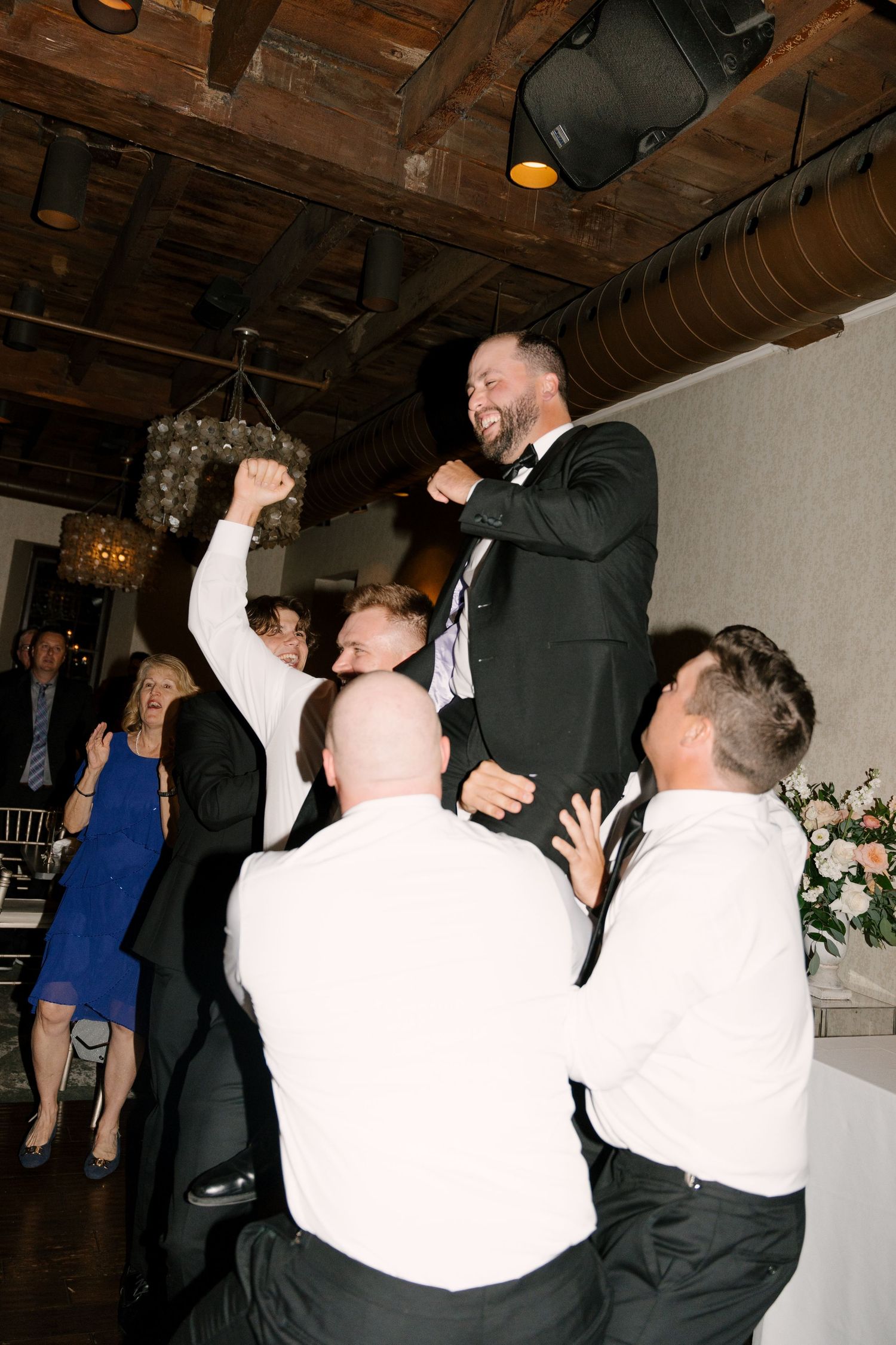 Groom in tuxedo is hoisted up by groomsmen during energetic wedding reception dance floor celebration. At Ancaster Mill venue.