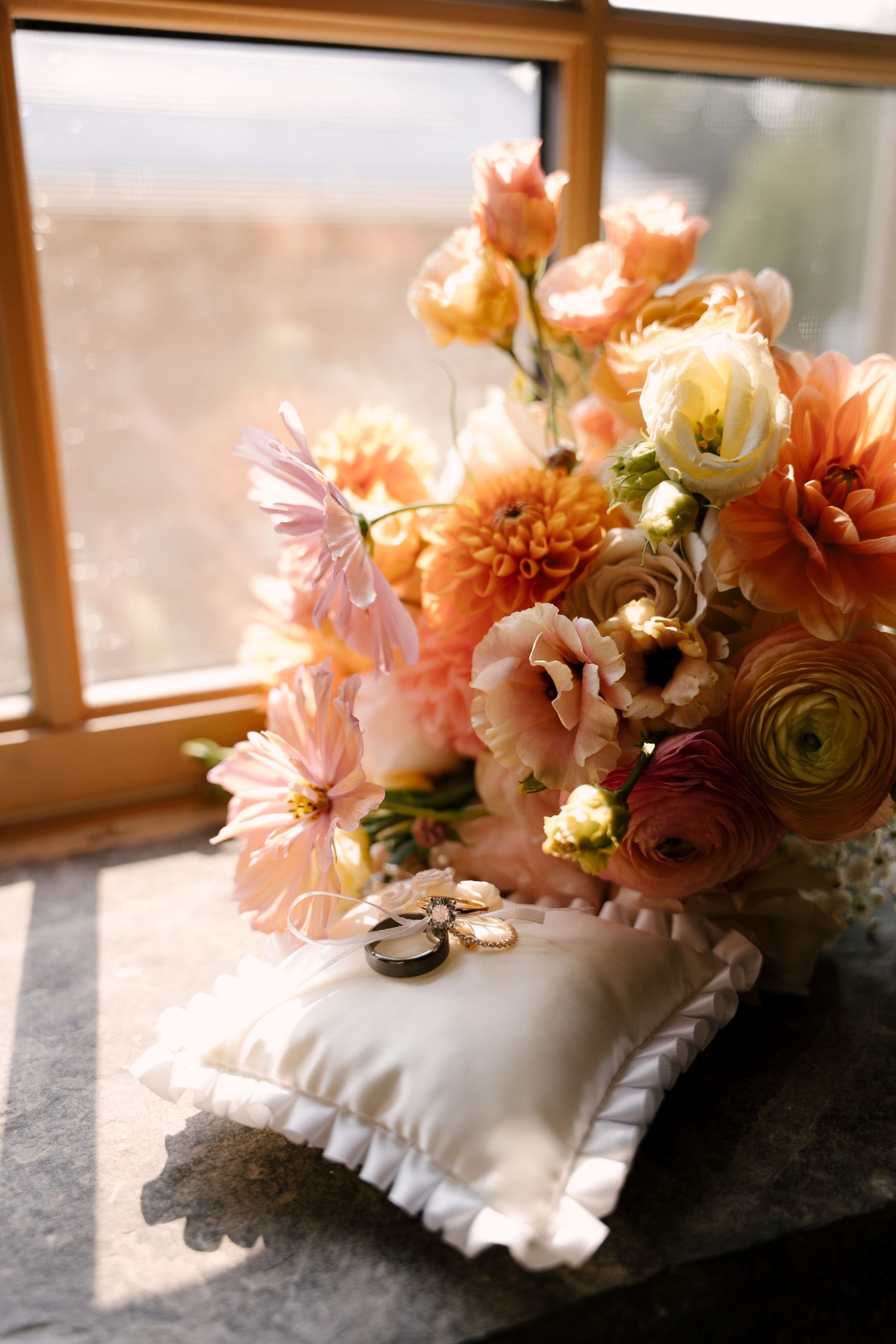 Wedding rings on white pillow beside peach dahlia and ranunculus bridal bouquet near sunlit window.
