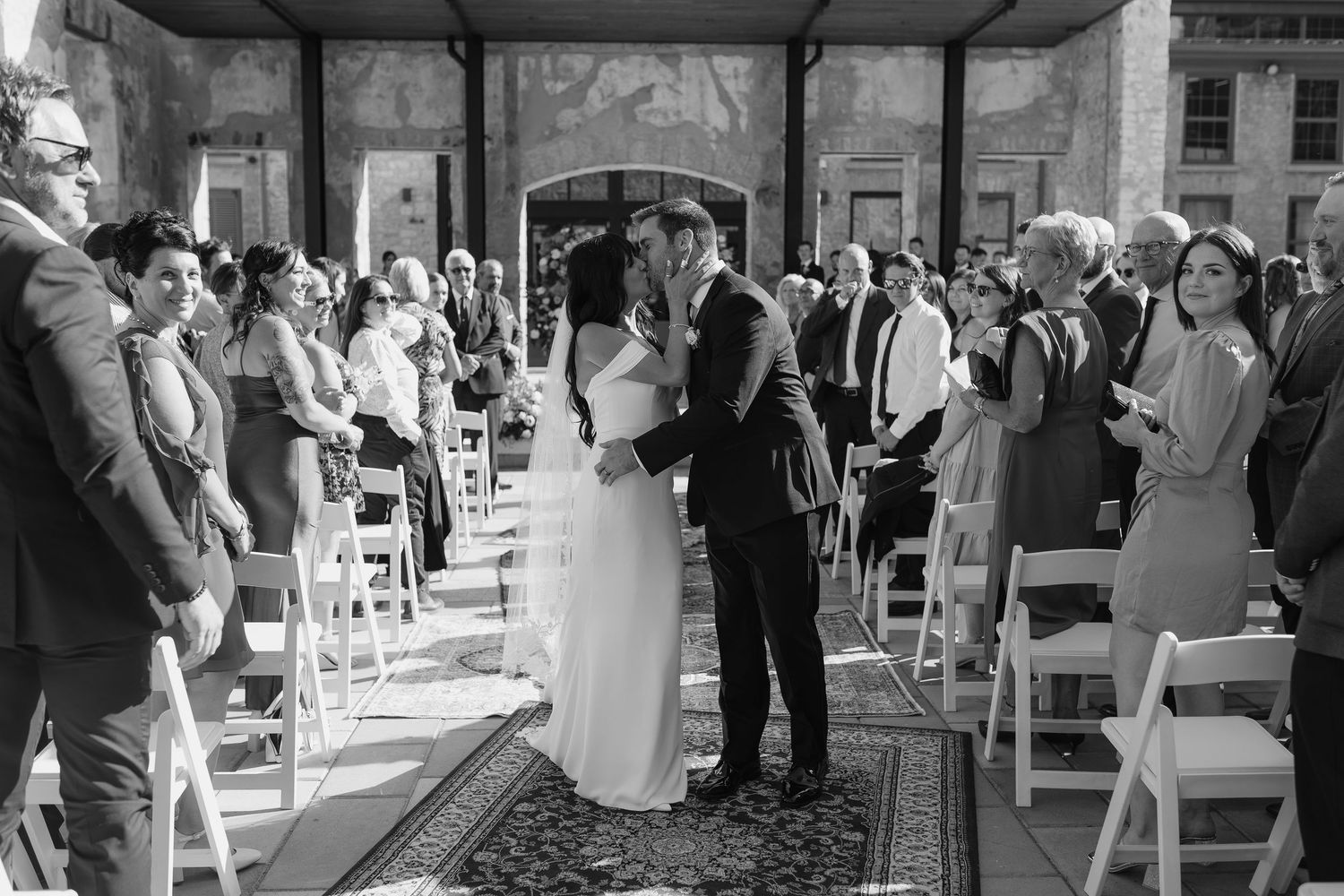 Black and white photo of bride and groom sharing first kiss at wedding ceremony aisle surrounded by guests. Outside Tapestry Hall venue.