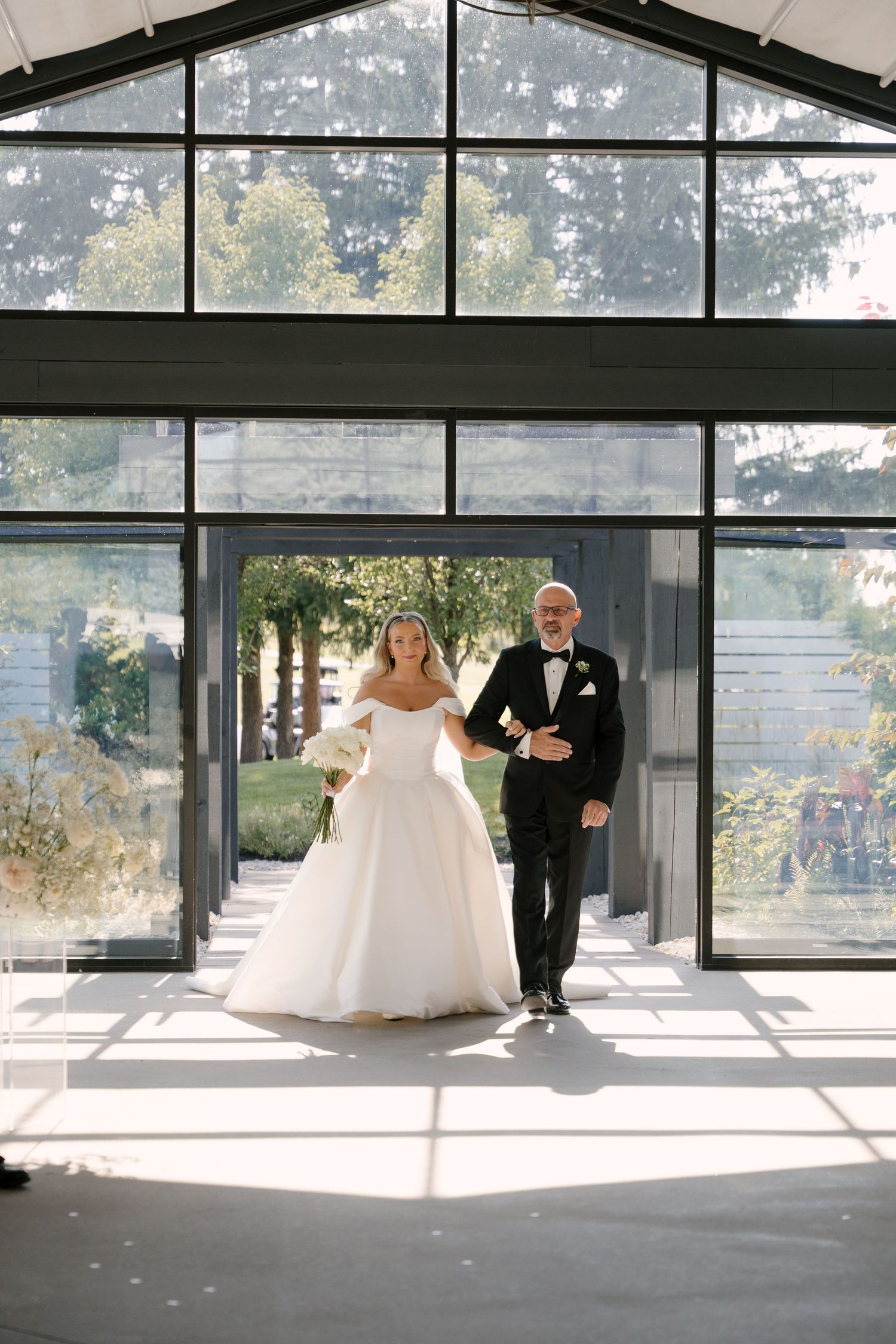 Bride in white ballgown entering glass greenhouse wedding venue with father escort.