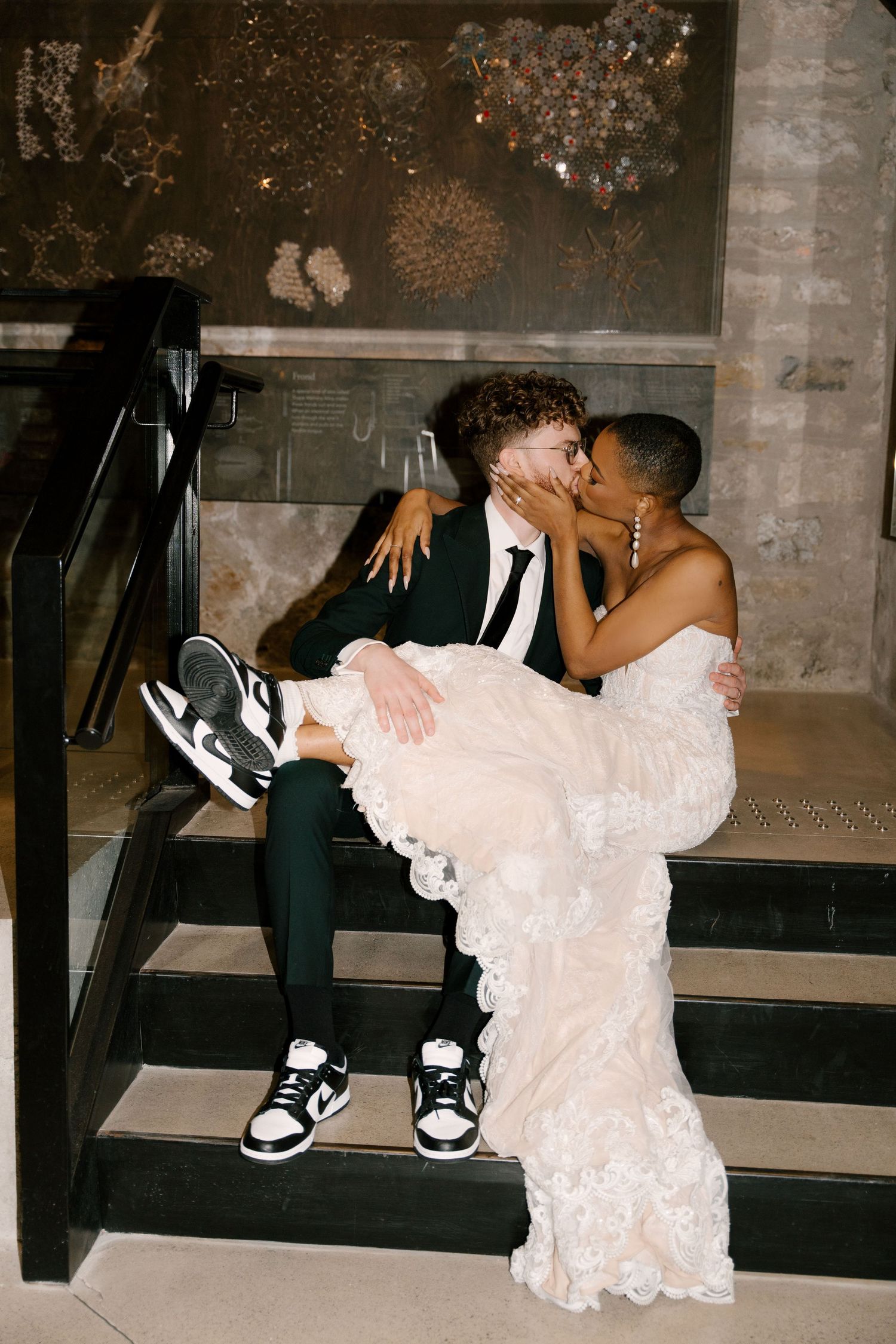 Newlywed couple sharing a romantic kiss on indoor stairs at Tapestry Hall, groom wearing Nike sneakers with bride in lace gown.