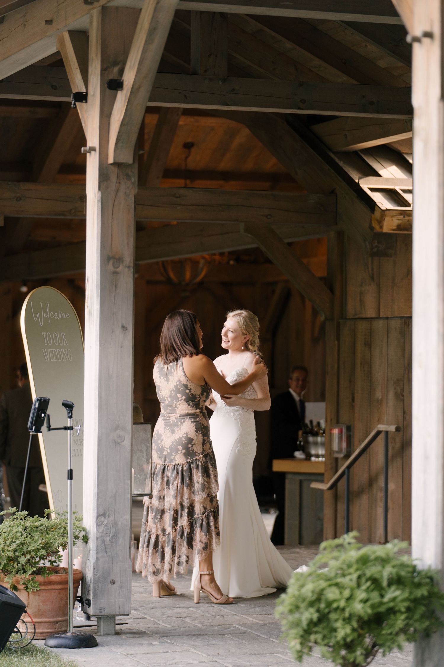 Bride in white gown embraces guest in floral dress under rustic wooden barn structure.