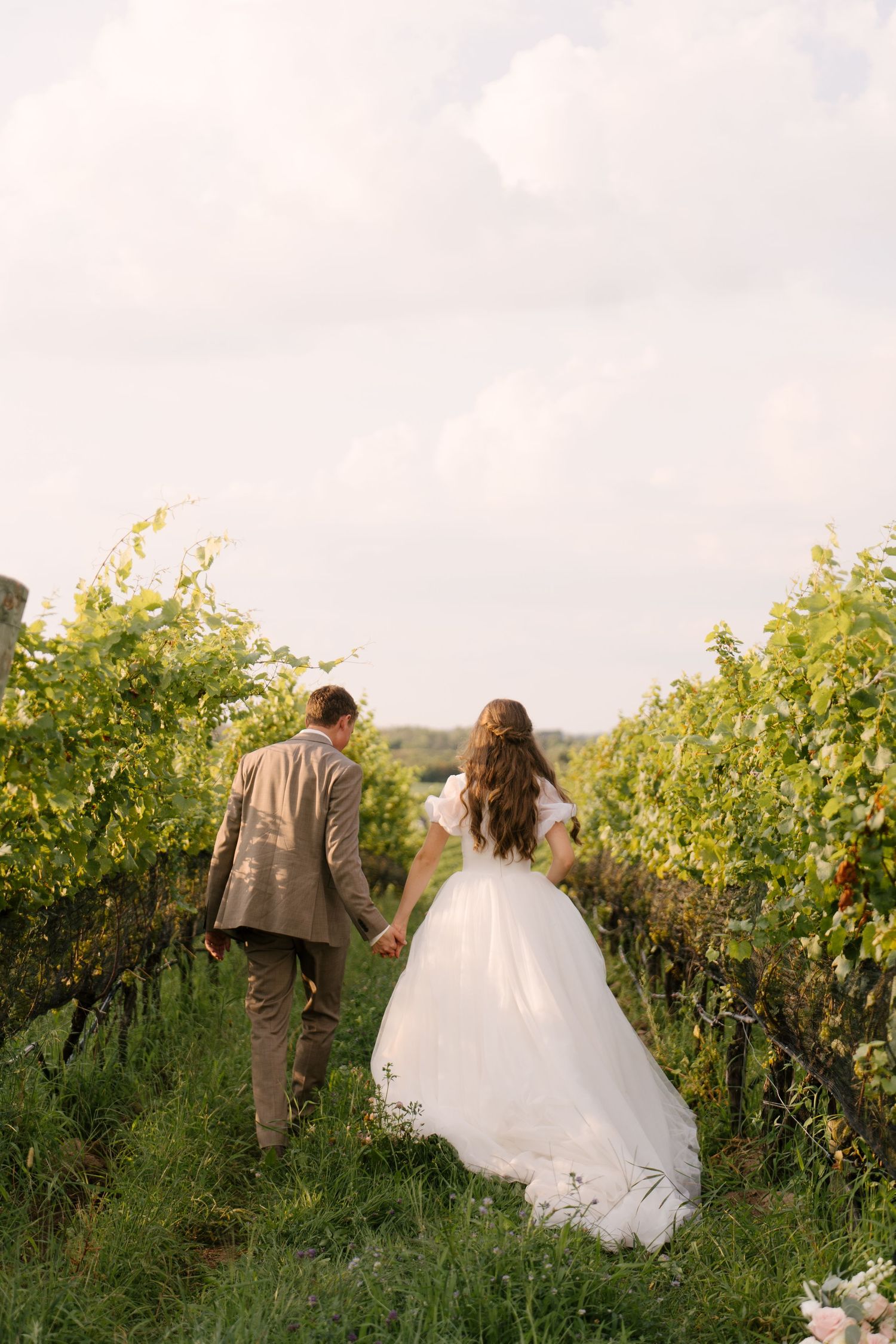 Bride and groom walking hand in hand through a scenic vineyard pathway on their wedding day.