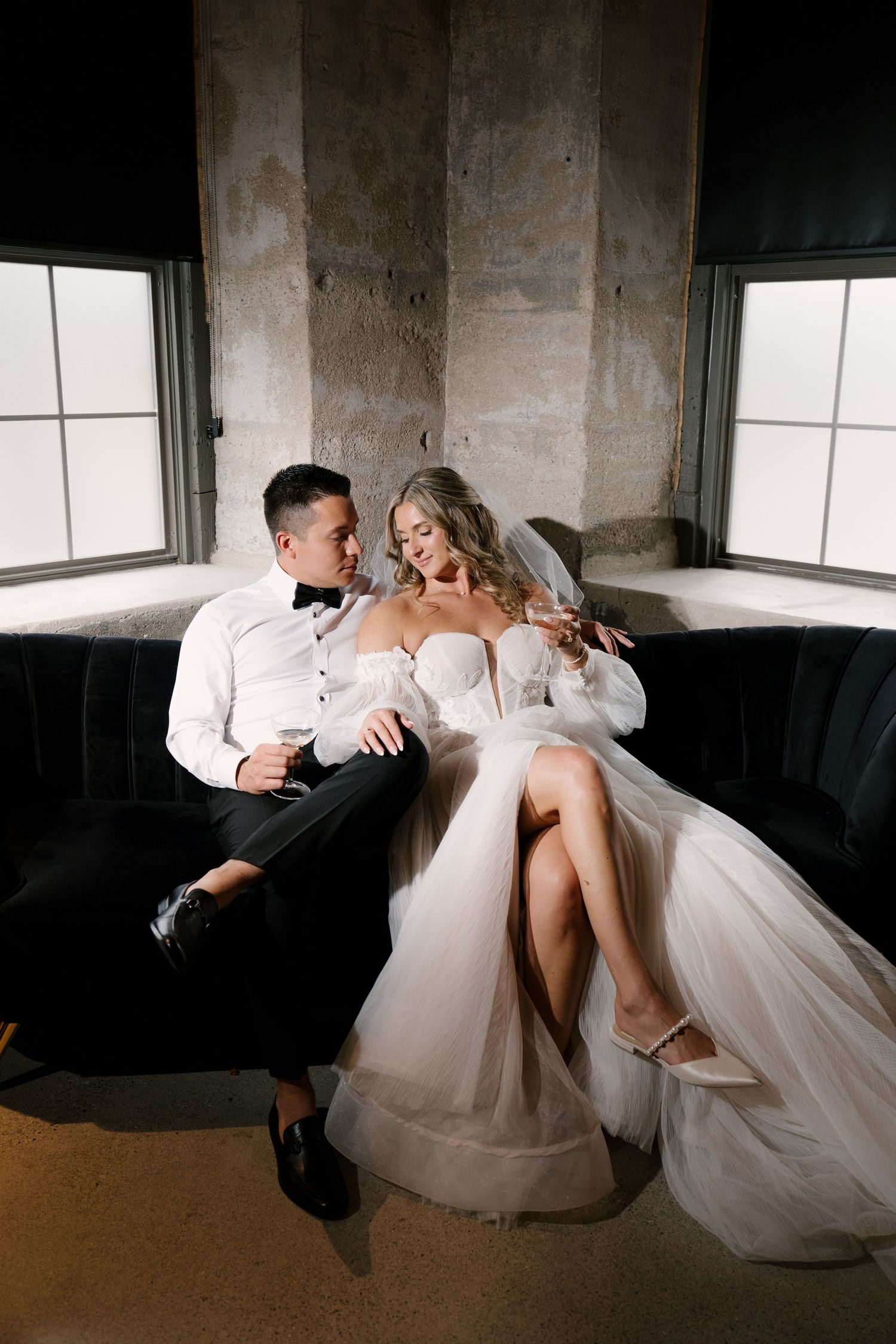 Bride and groom sitting intimately on a dark velvet sofa holding champagne glasses at Tapestry Hall.