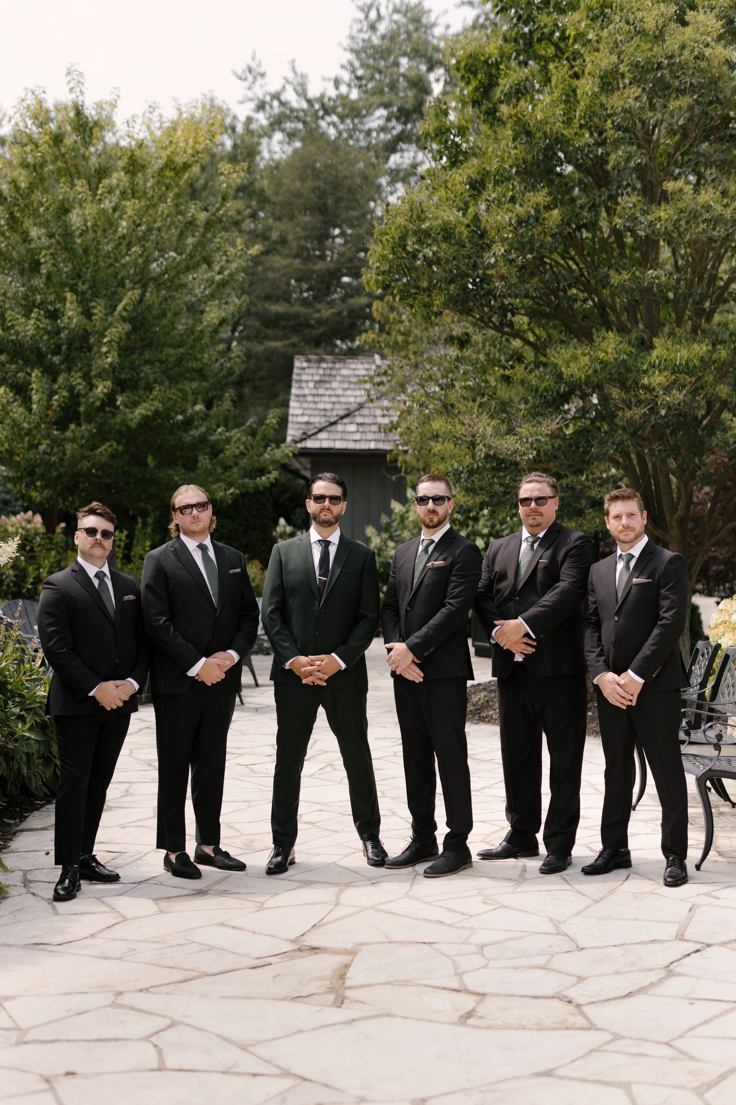Six groomsmen in black suits and sunglasses pose on a stone patio surrounded by lush green trees at Roseville Estates wedding venue.