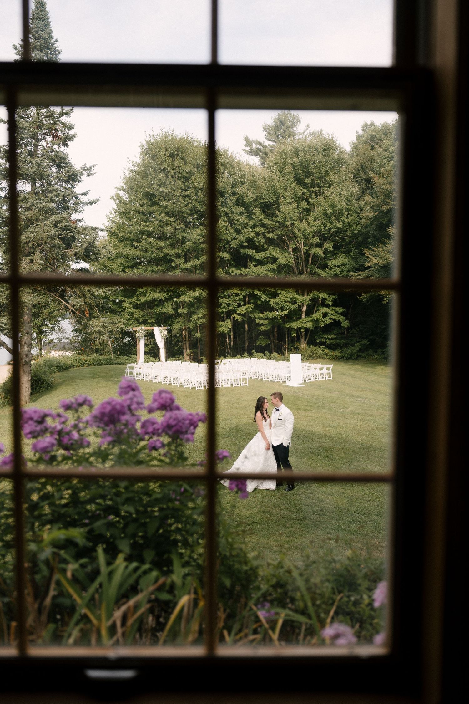 Bride and groom embrace near a white ceremony arch on a green lawn, viewed through a vintage window frame.