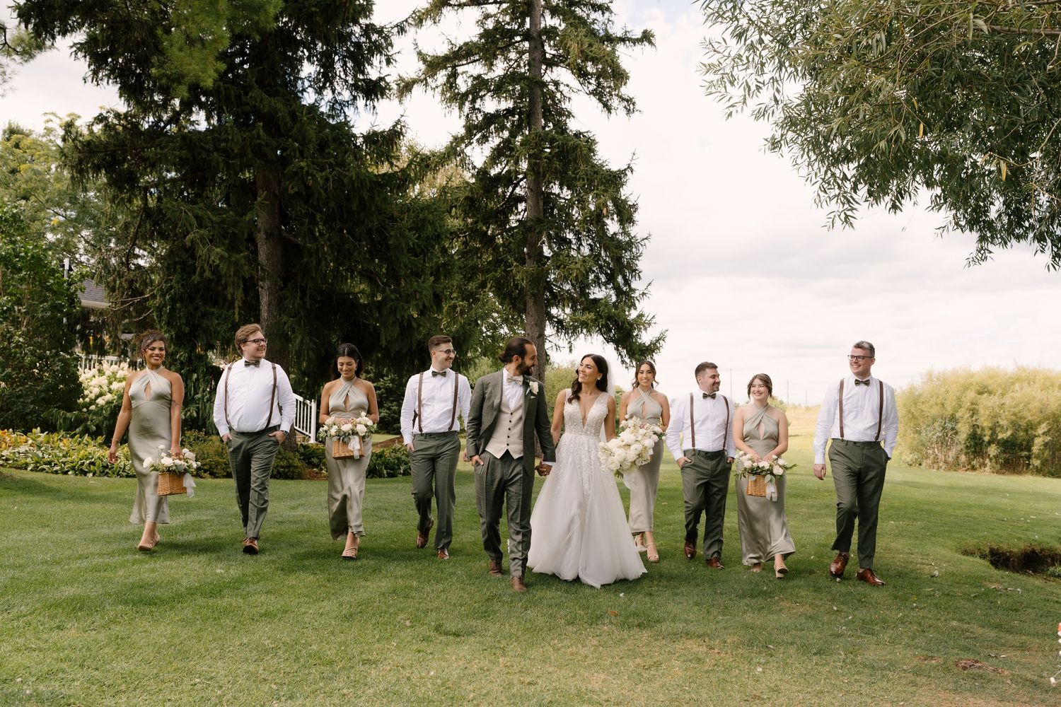 Wedding party walks together on green lawn, bridesmaids in sage dresses and groomsmen in gray suits with suspenders.