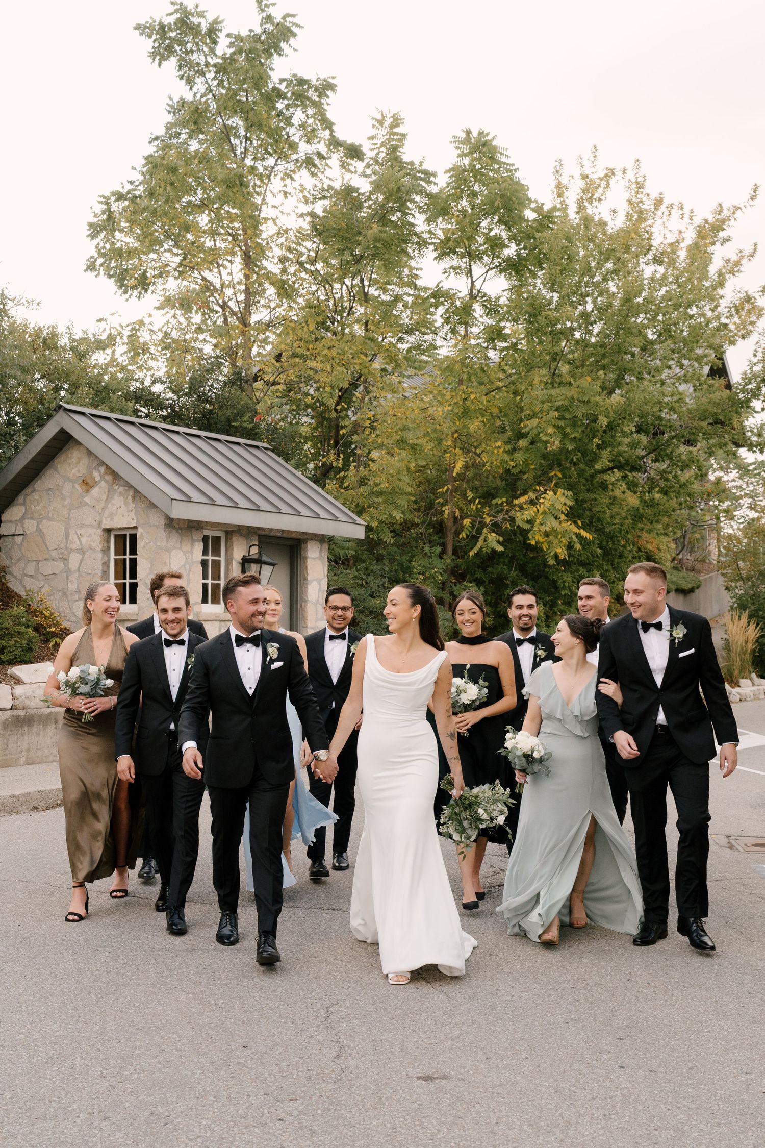 Bride and groom walk hand-in-hand with smiling wedding party on a tree-lined outdoor path at Cambridge Mill.