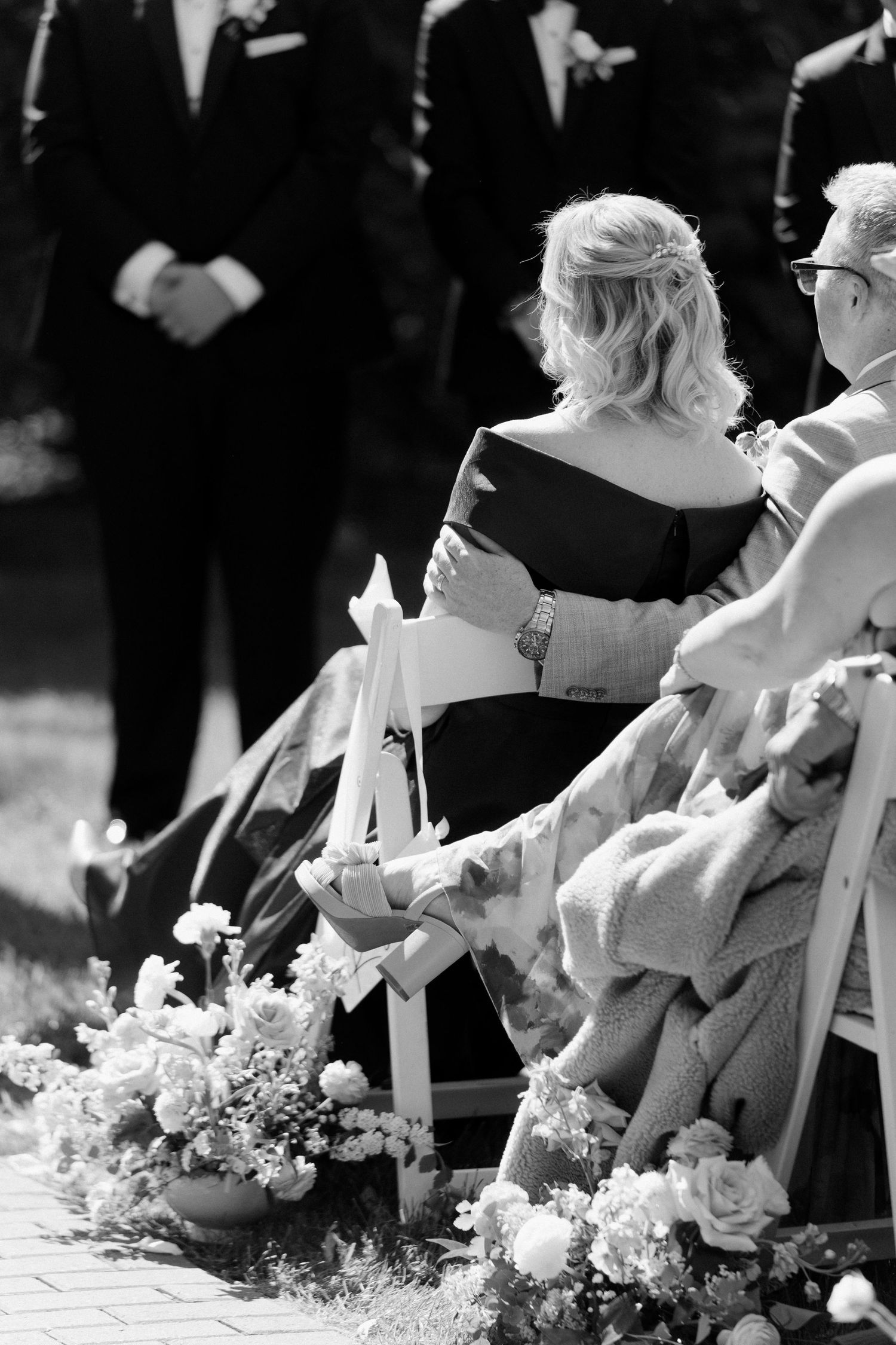 Wedding guest in black off-shoulder dress seated at floral-decorated outdoor ceremony.
