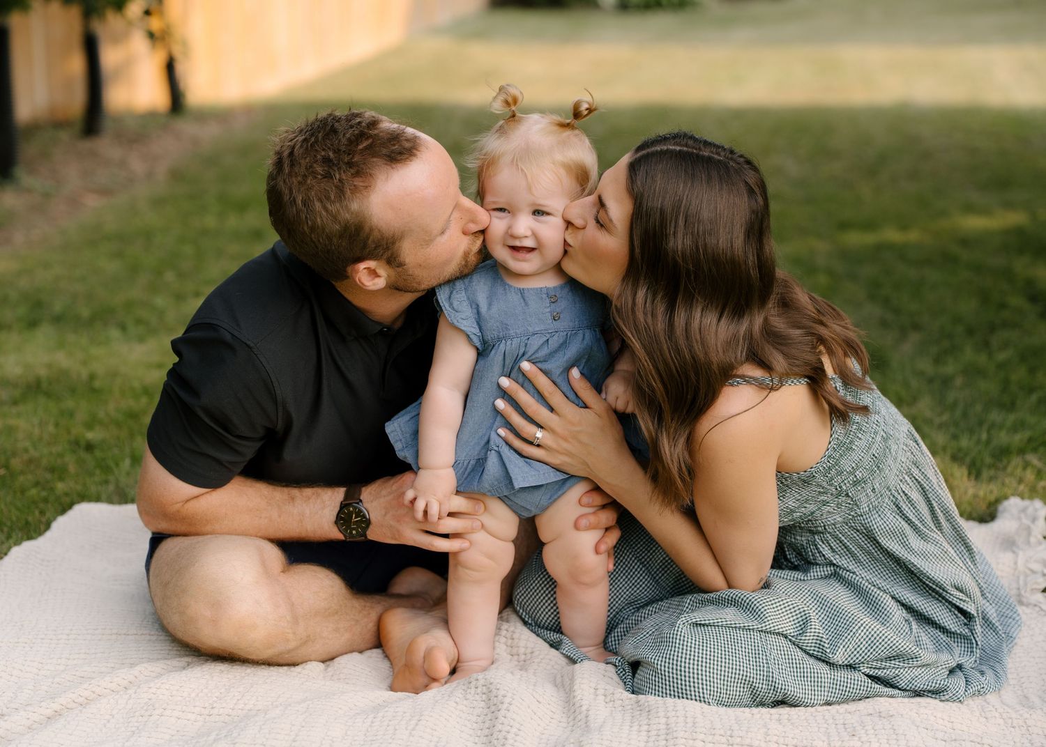 Loving parents kiss their smiling toddler daughter on the cheeks while sitting on a blanket outdoors.