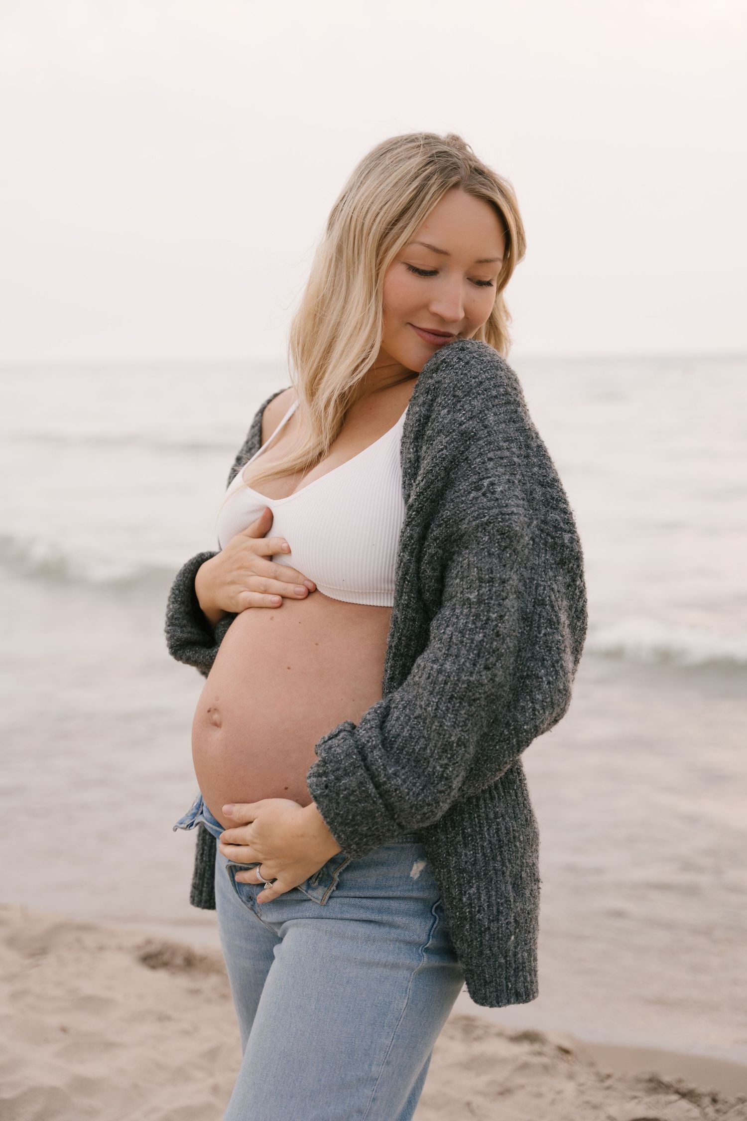 Glowing pregnant woman in gray cardigan lovingly gazing down at her bare baby bump on beach.