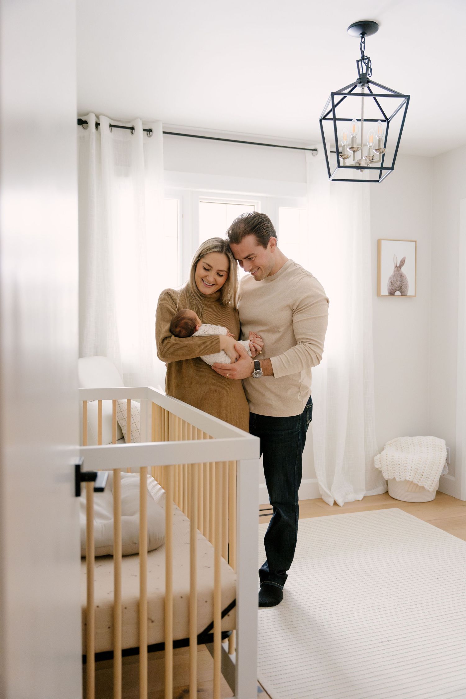 Smiling couple admiring newborn baby together in bright modern nursery with white crib and pendant light.