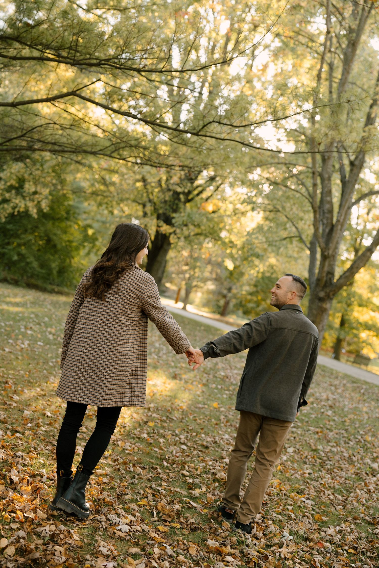 Couple holding hands walking through leaf-covered autumn park, man smiling back at woman.