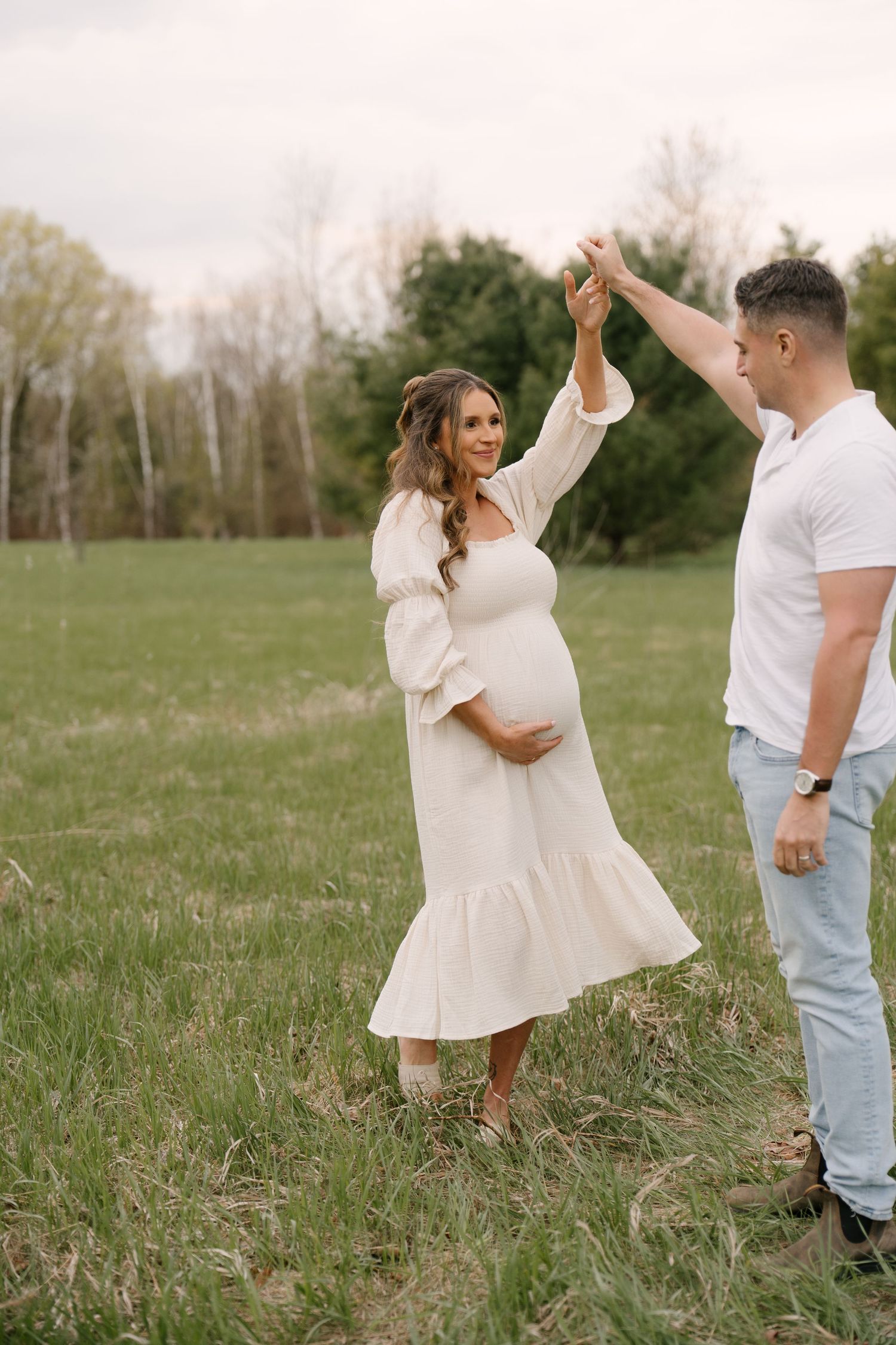 Happy pregnant couple dancing together in a lush green meadow at sunset.