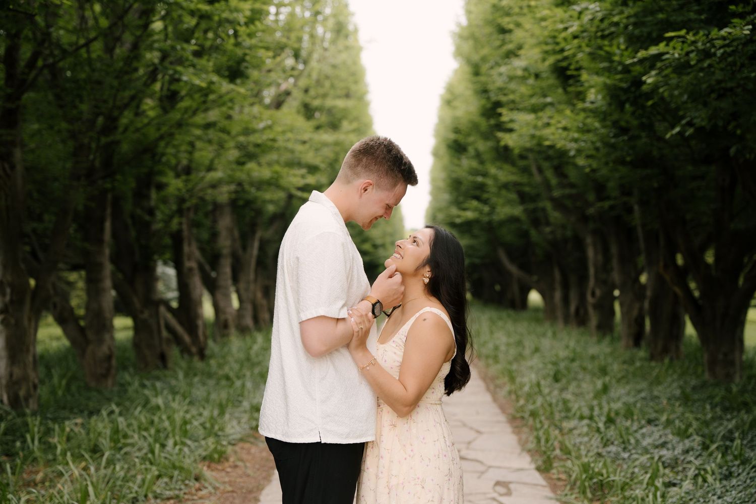 Romantic couple facing each other on a tree-lined path with soft green foliage surroundings.