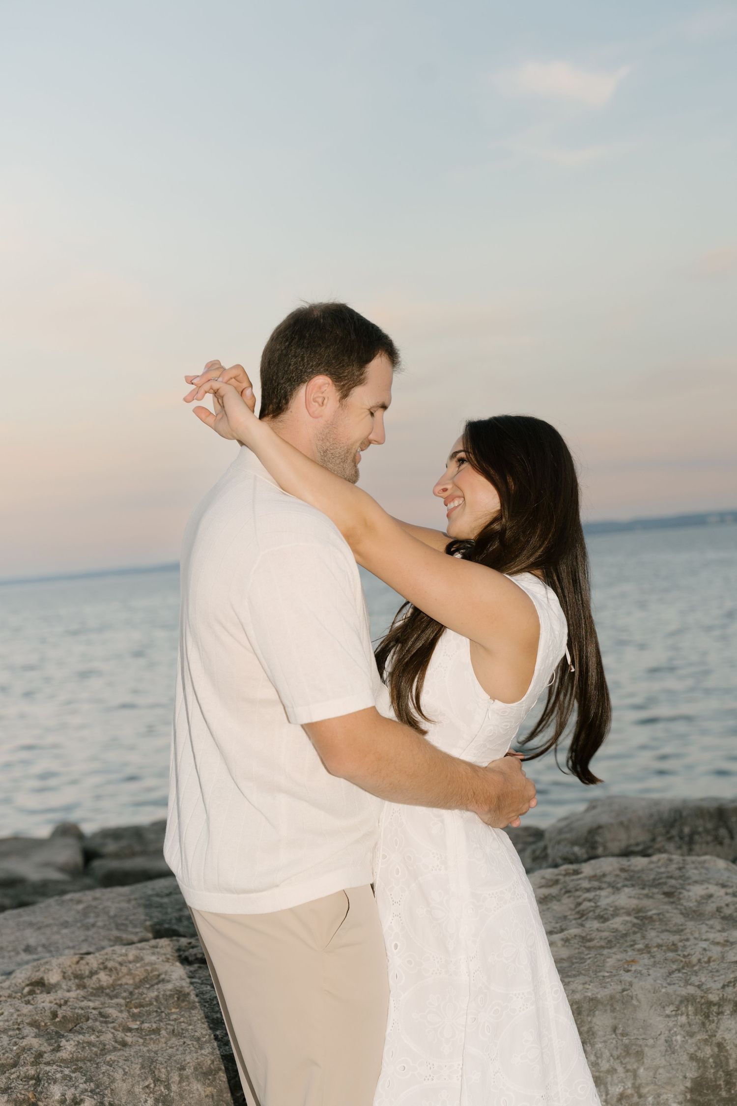 Smiling couple embracing on rocky shoreline at dusk, woman in white dress with ocean backdrop.