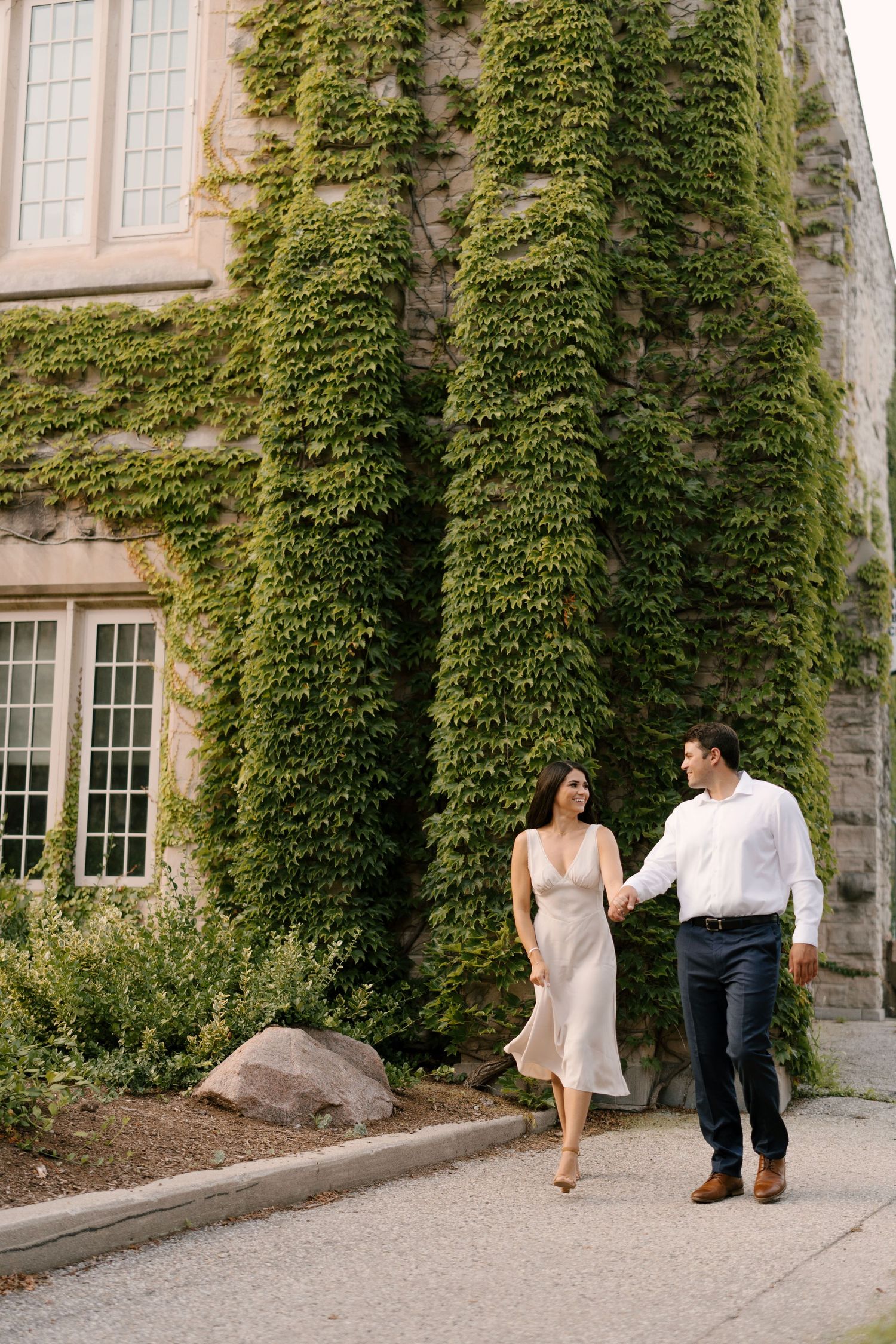 Engaged couple walking hand in hand past ivy-covered stone building in warm summer light.