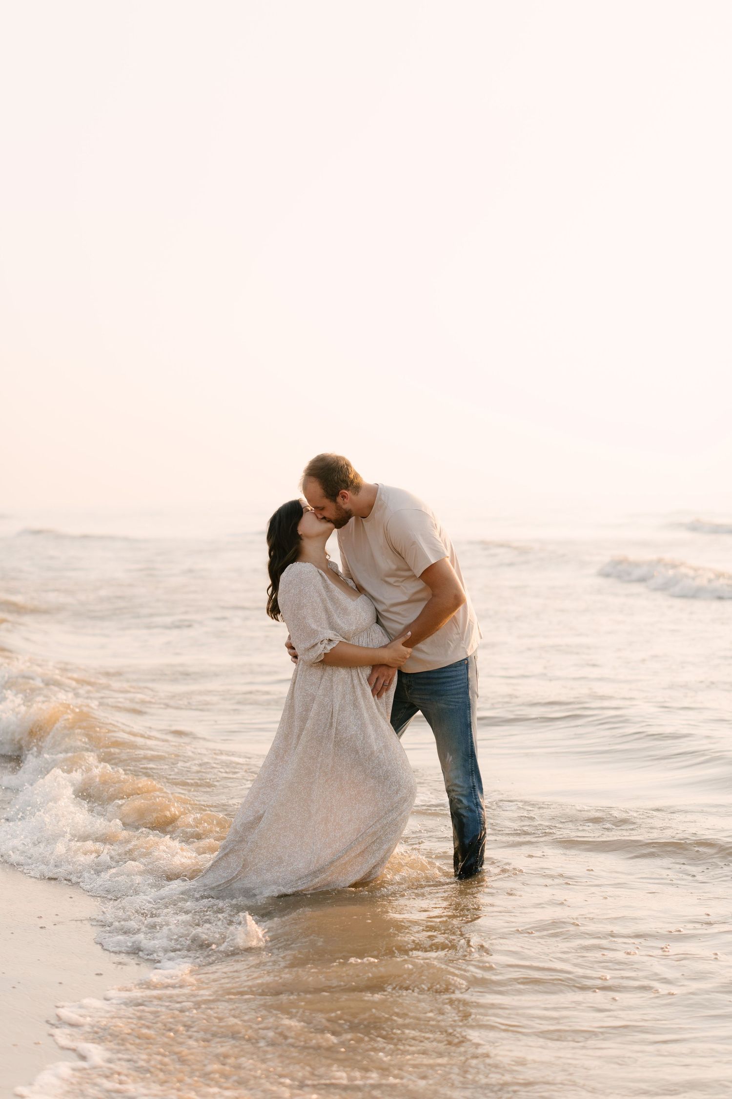 Couple sharing romantic kiss in ocean waves at sunset, pregnant woman in flowing white dress.