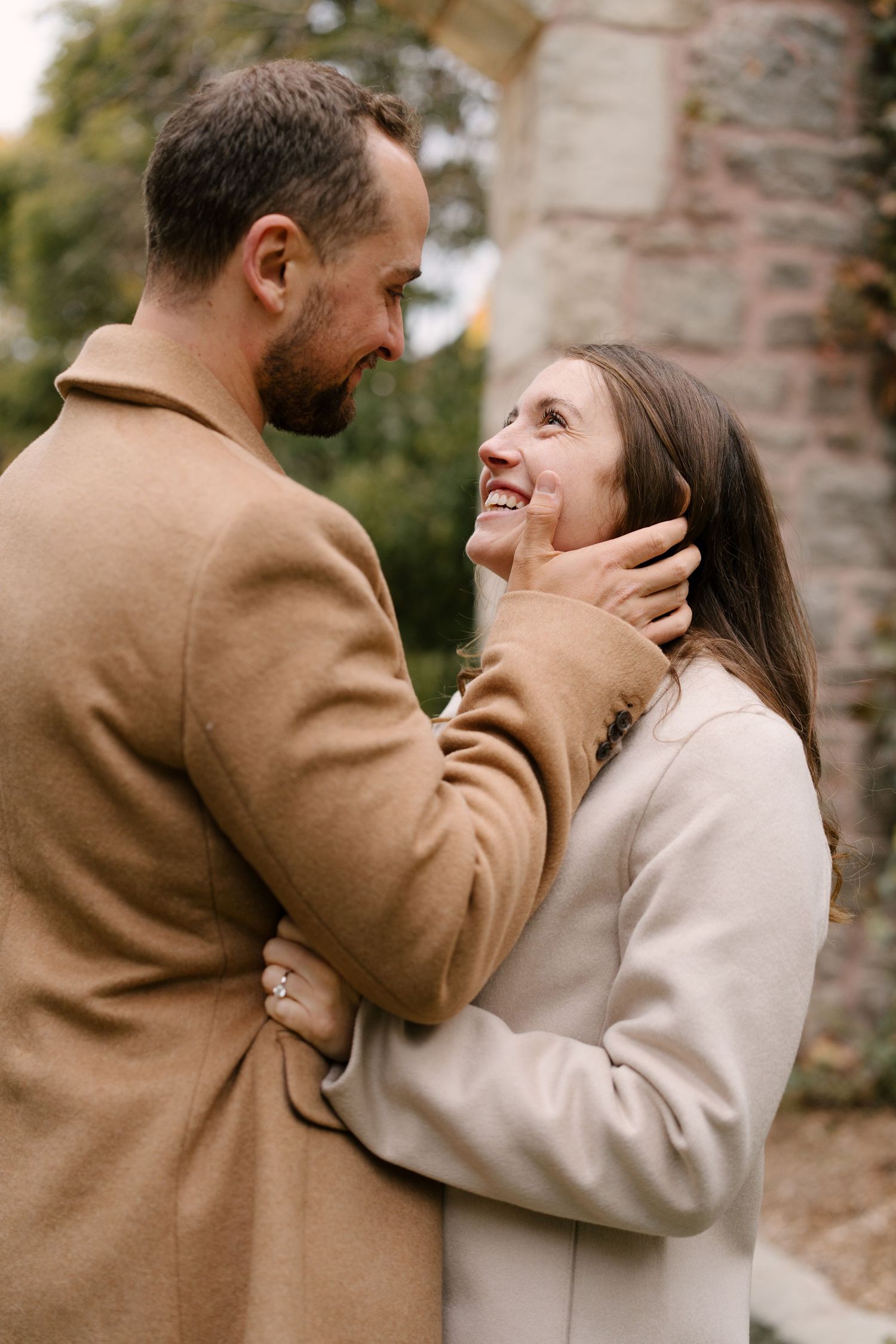 Man tenderly cupping smiling woman's face as they gaze at each other, both in beige wool coats.