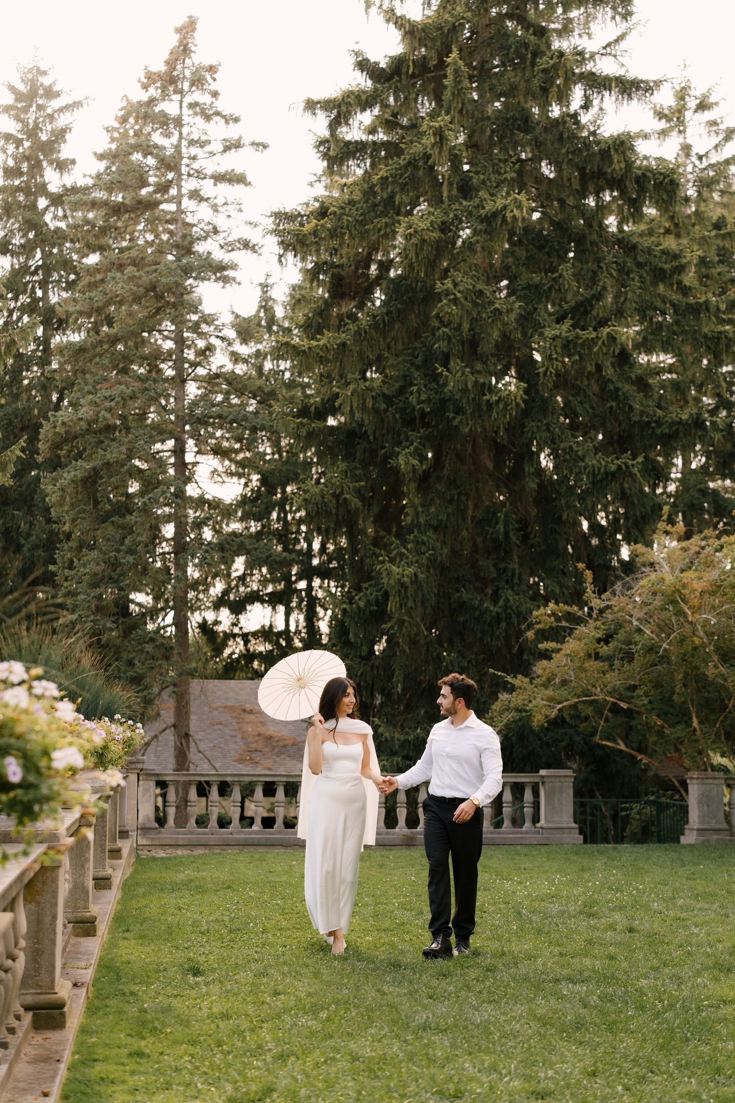 Bride in white dress holding parasol walks hand-in-hand with groom across manicured lawn beside stone balustrade.