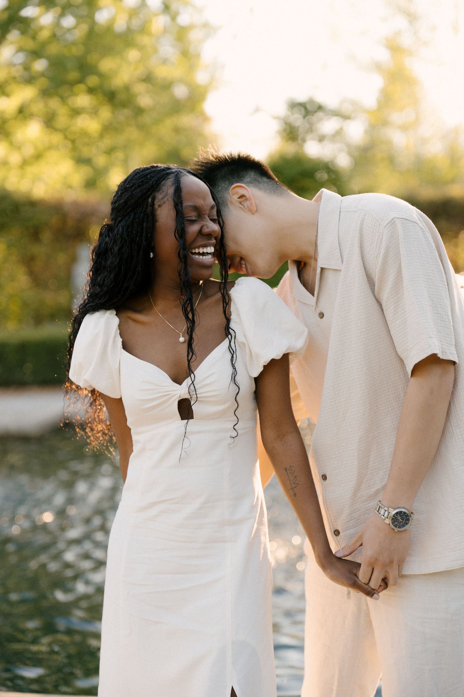 Laughing couple holding hands near a reflective pond in a sun-drenched park at golden hour.