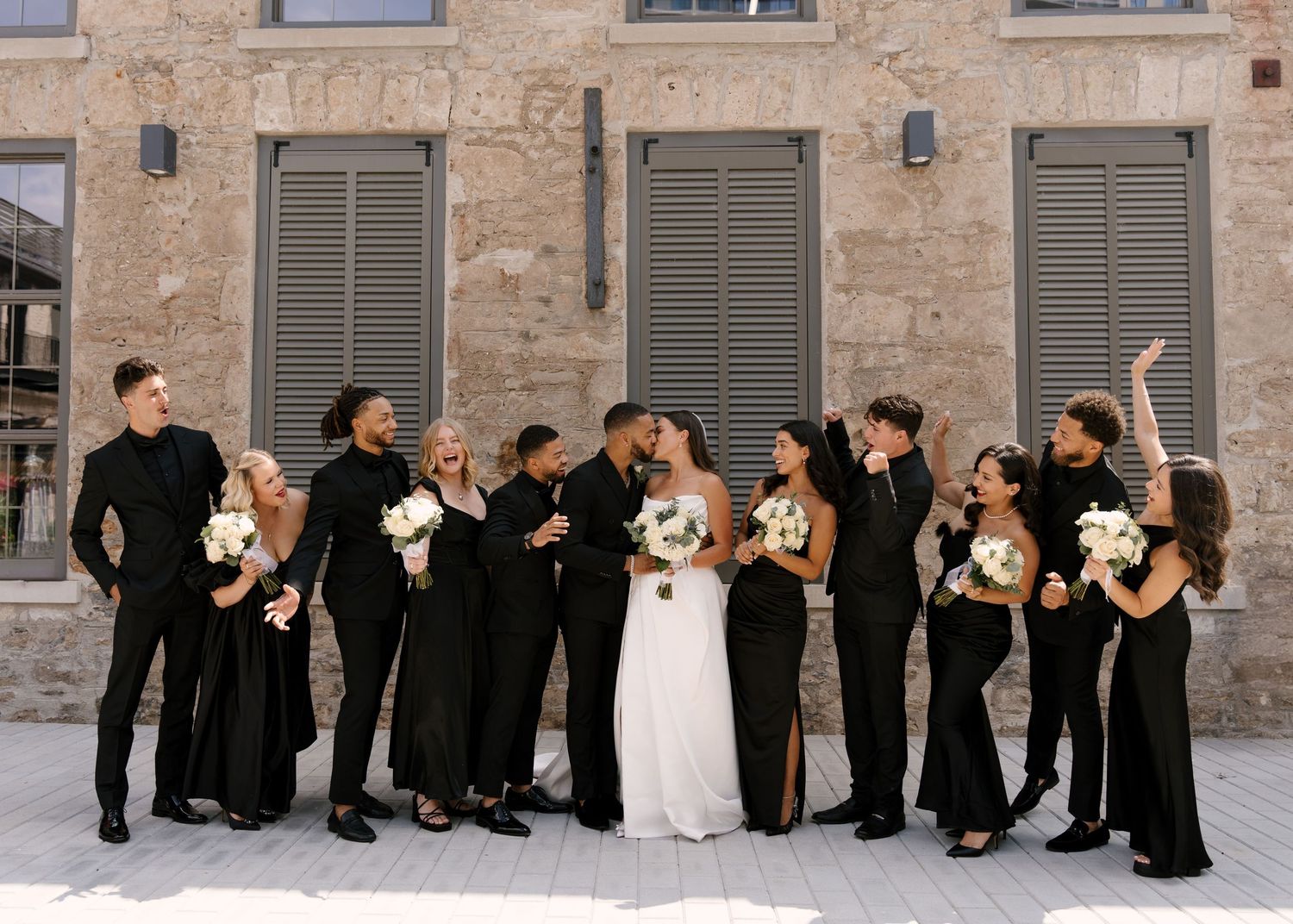 Wedding party in all-black attire with bride in white gown holding bouquet, posing outdoors near stone building.