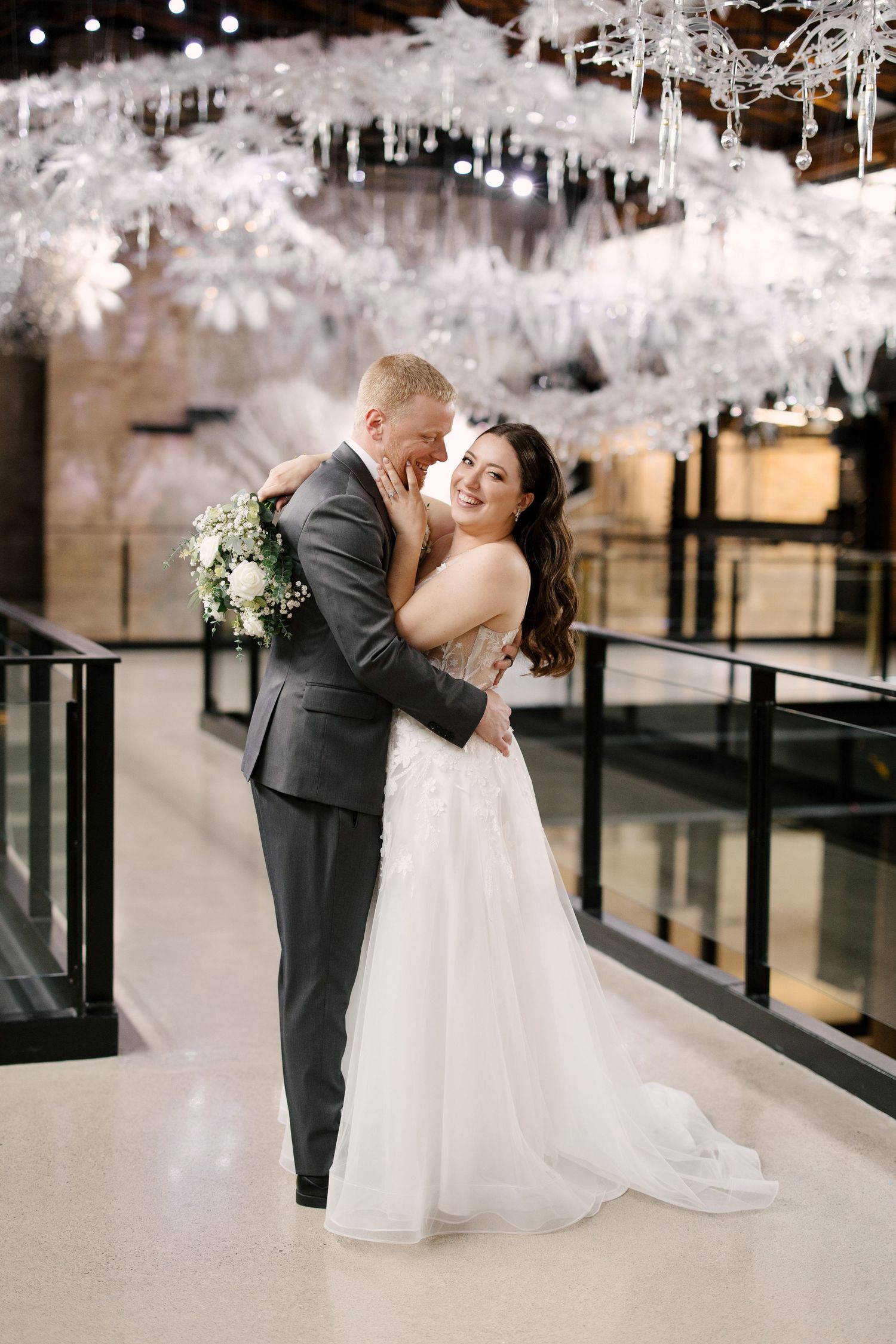 Bride and groom embracing under white floral chandelier installation in a modern industrial wedding venue.