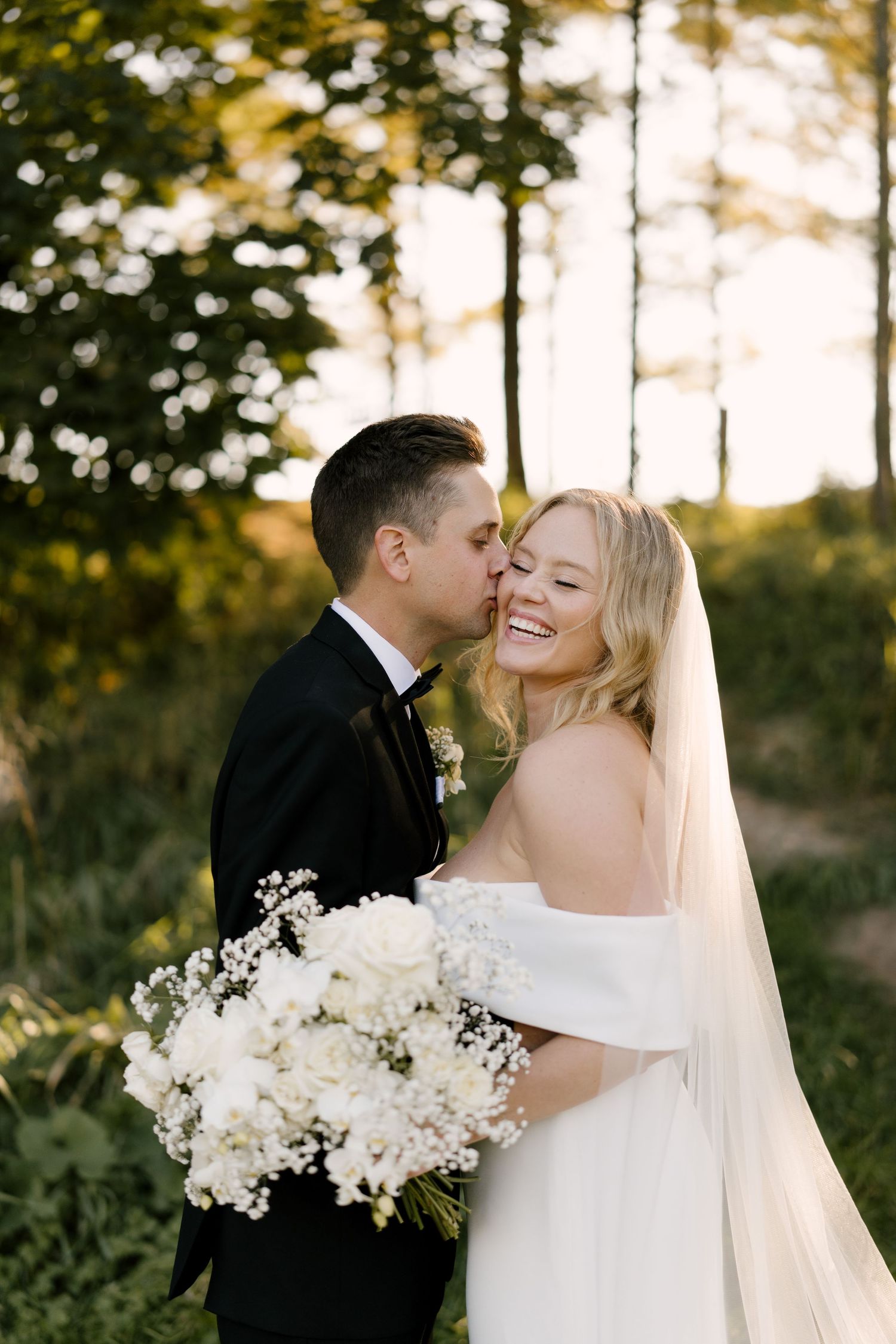 Bride laughing joyfully as groom kisses her cheek outdoors, holding white floral bouquet at golden hour at Cellar 52.