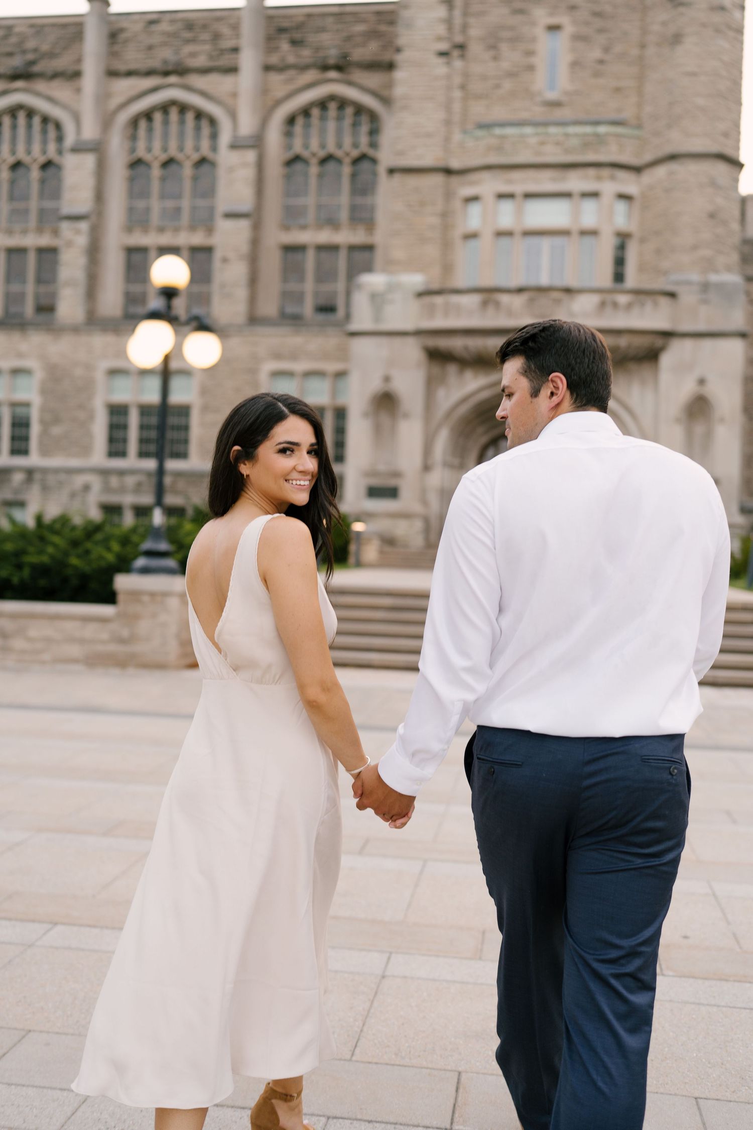 Woman in white dress smiles back at camera while holding hands with man near stone building.