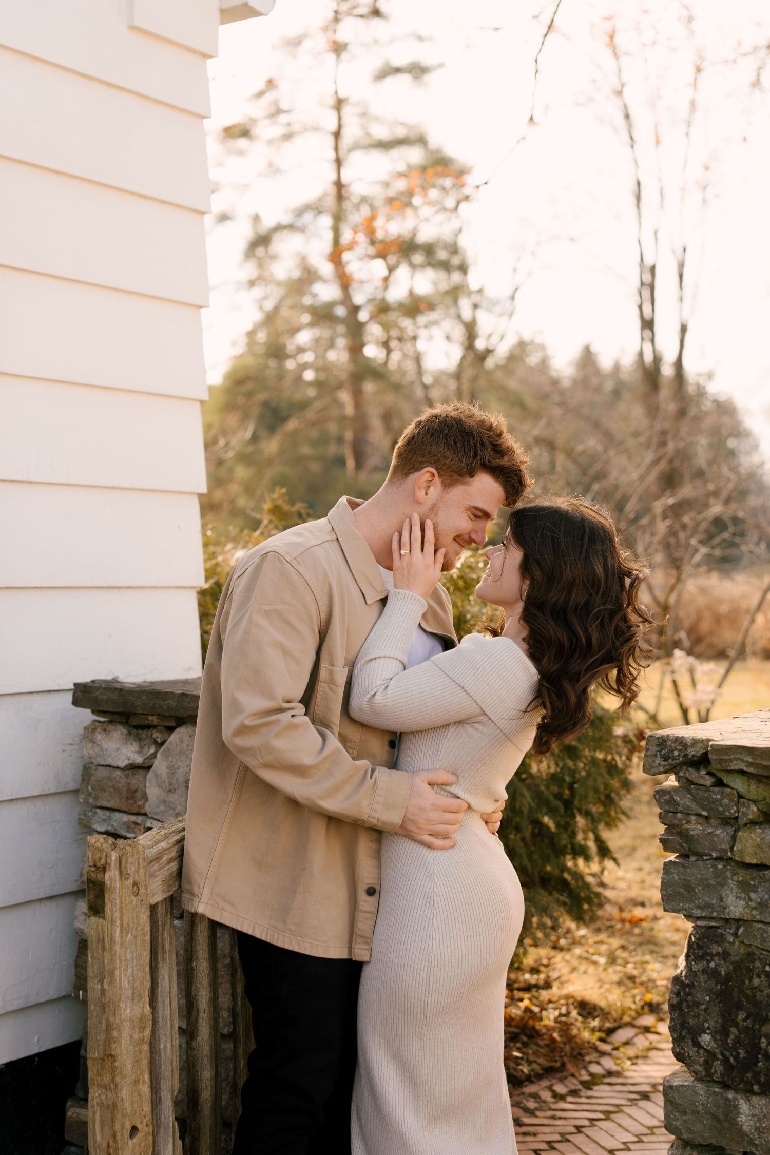 Couple sharing a tender kiss beside a white building with stone foundation at golden hour.