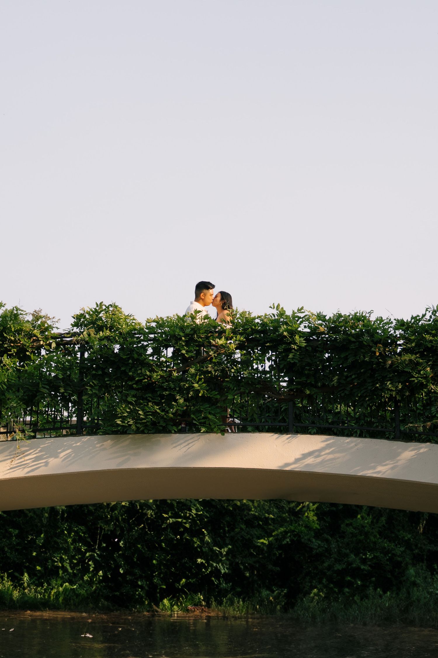 Couple sharing a kiss on an arched bridge overlooking a tranquil green waterway at sunset.