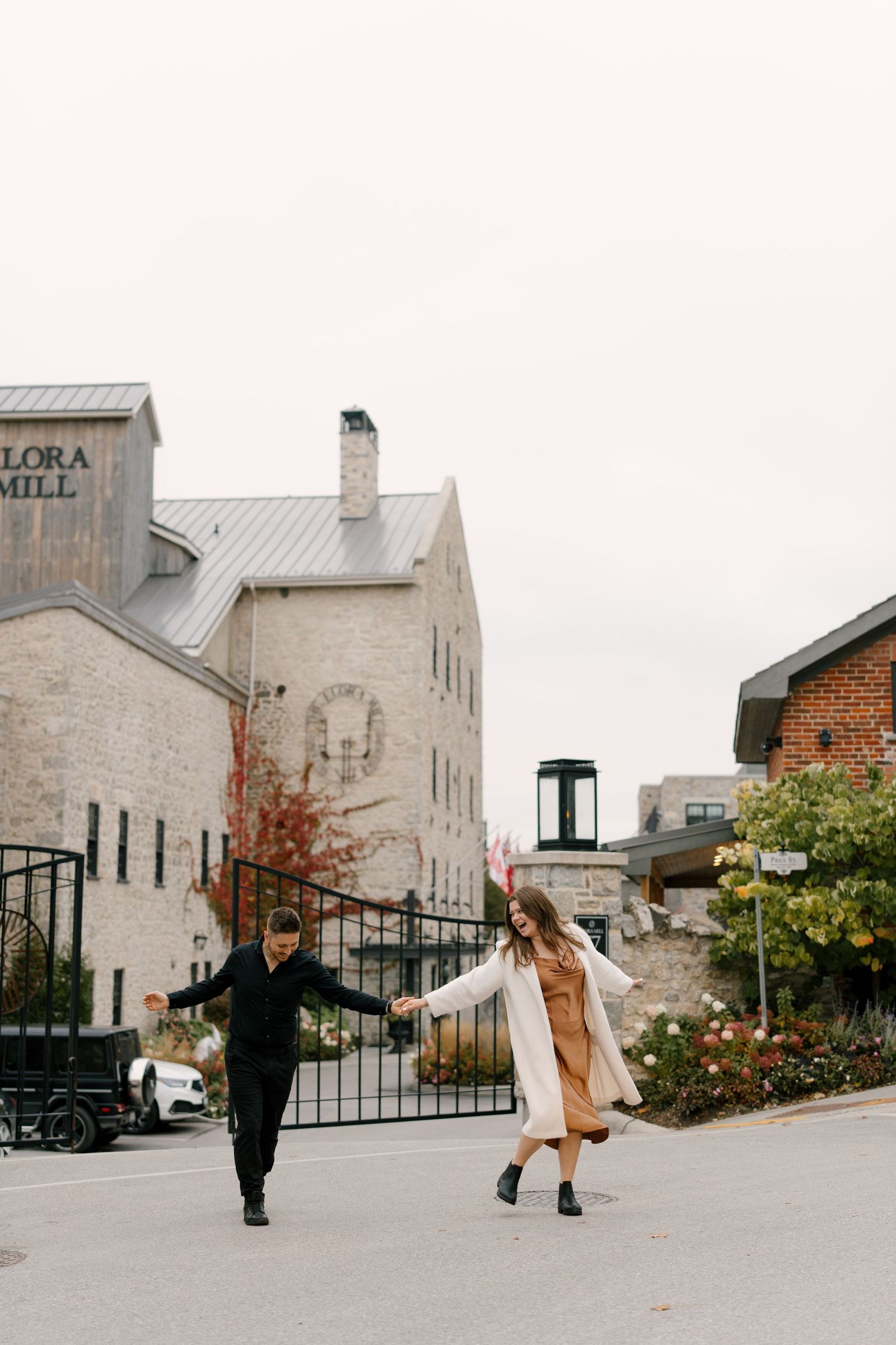 Couple walking hand in hand in front of the historic Elora Mill building on an overcast autumn day.