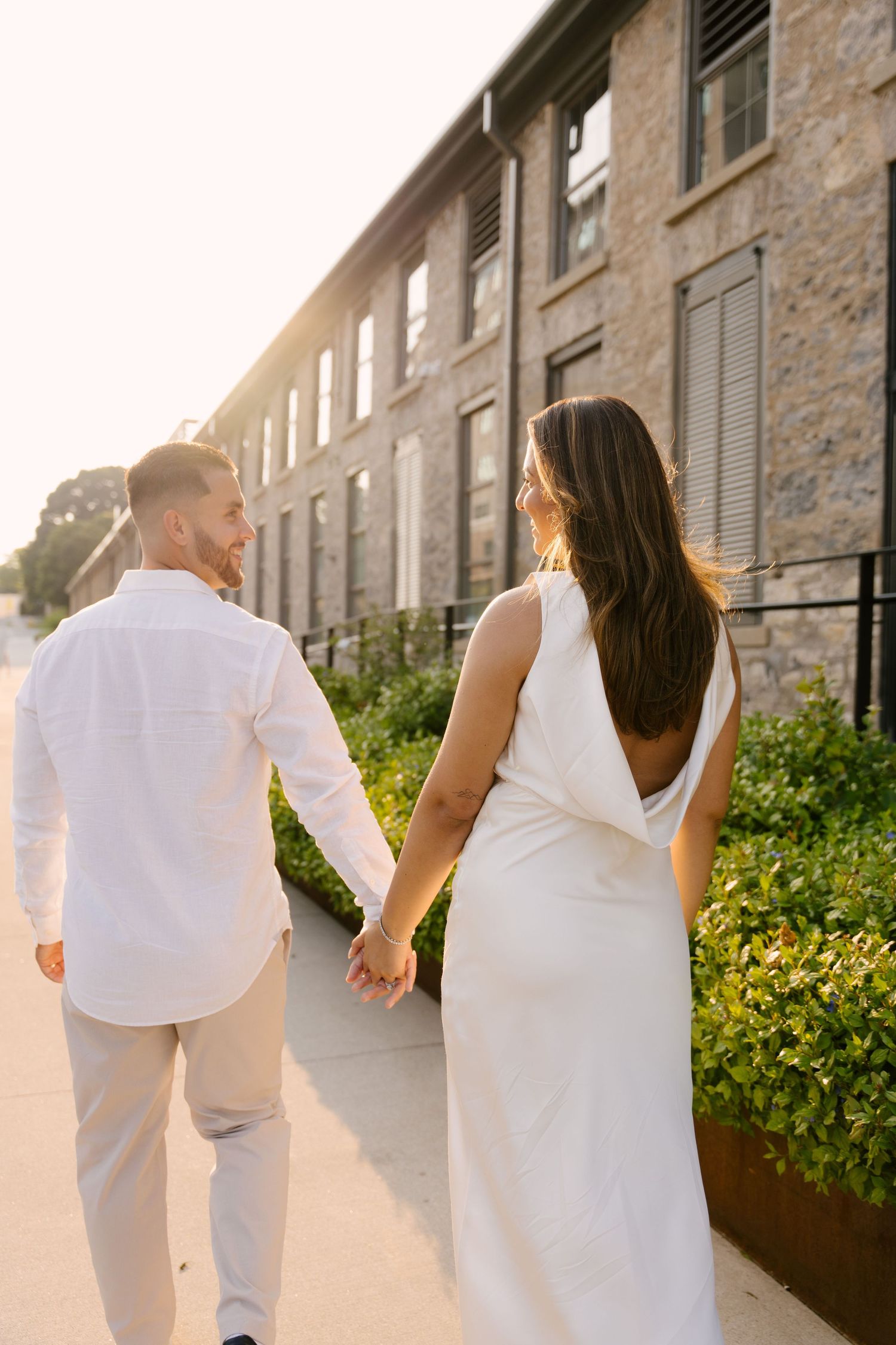 A couple walks hand-in-hand past a historic stone building at golden hour, the woman in a white backless dress.