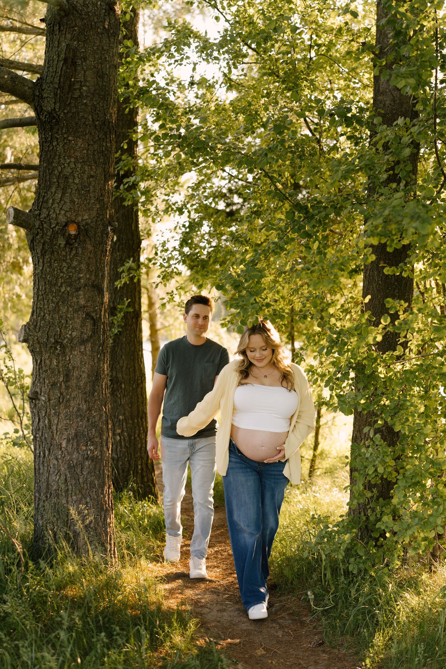 Pregnant woman in yellow cardigan walks a forest path with her partner during a golden hour maternity shoot.