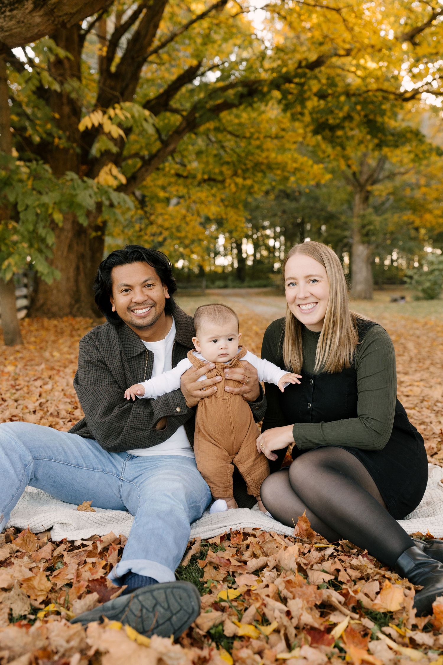 Smiling interracial family sitting on autumn leaves in park, parents with baby between them.