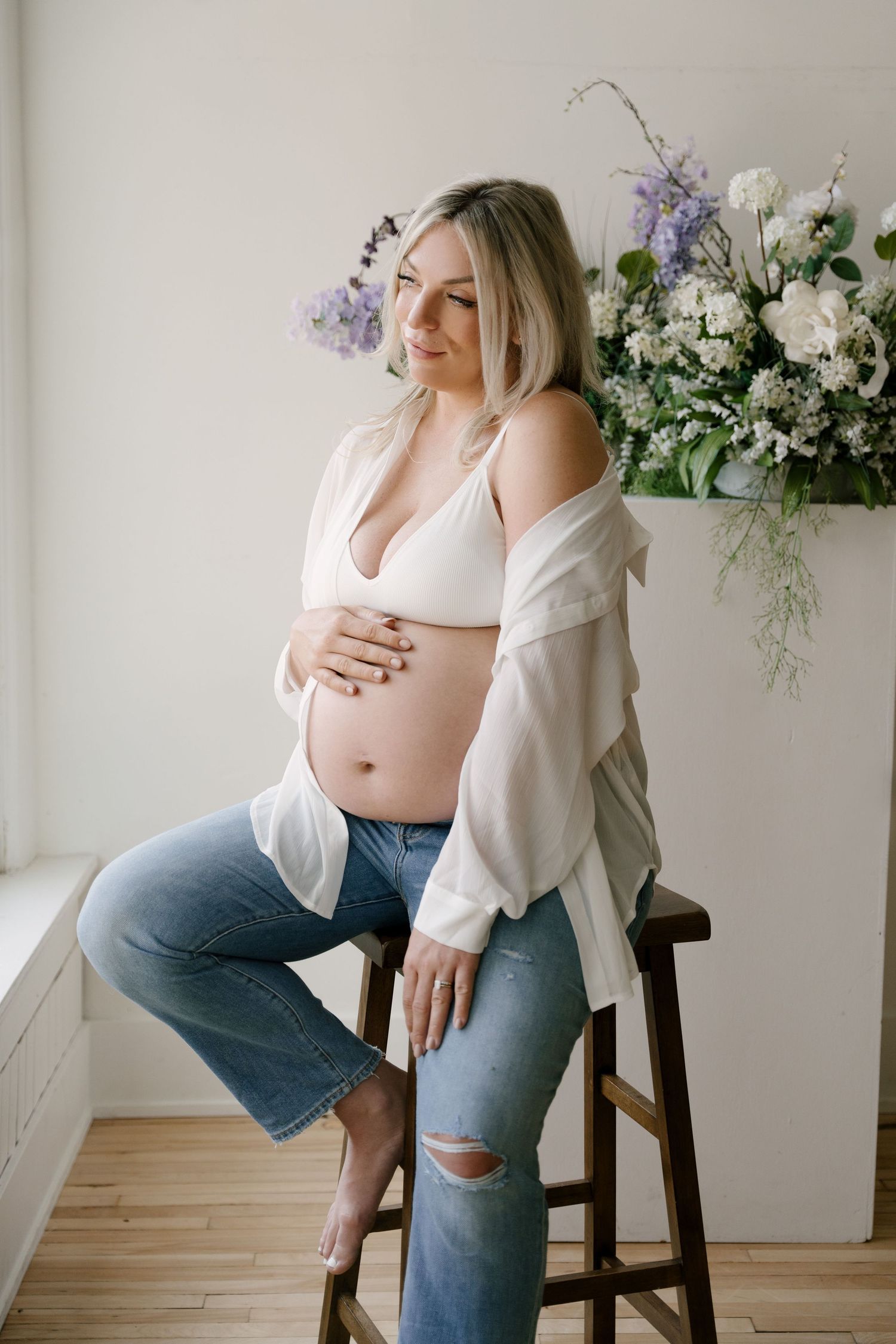 Pregnant woman in white bralette and ripped jeans sits on stool, cradling baby bump near floral arrangement.