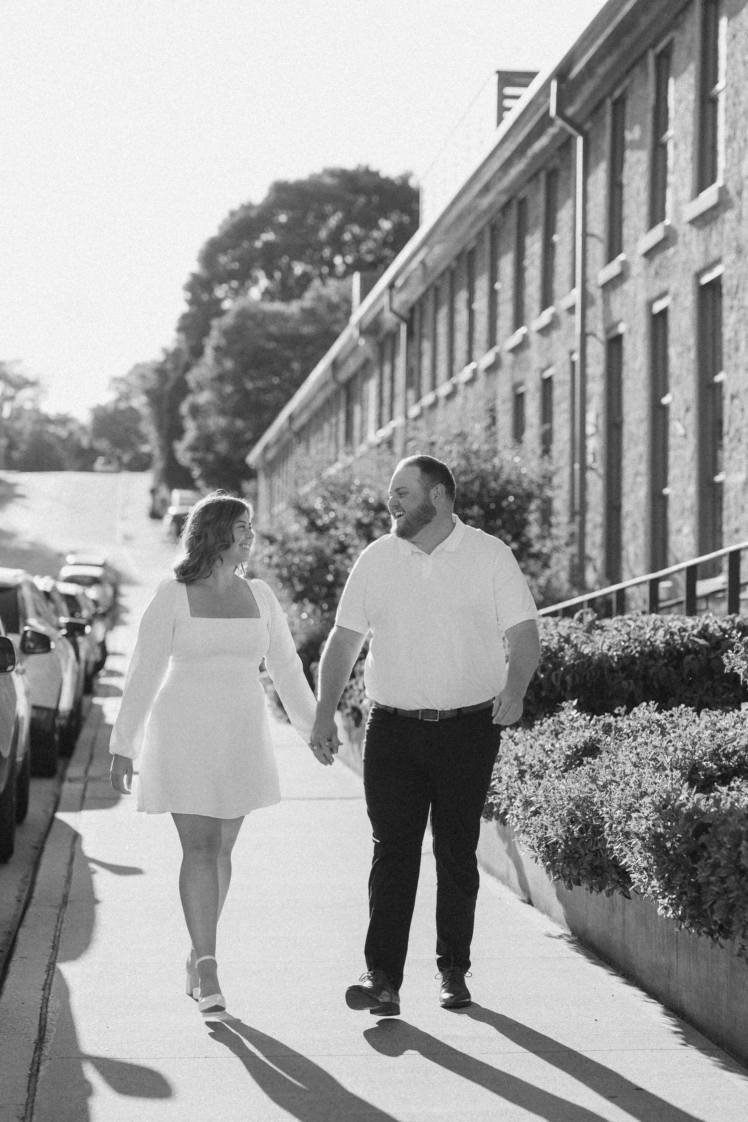 Black and white photo of couple holding hands and gazing at each other on a sunny sidewalk.