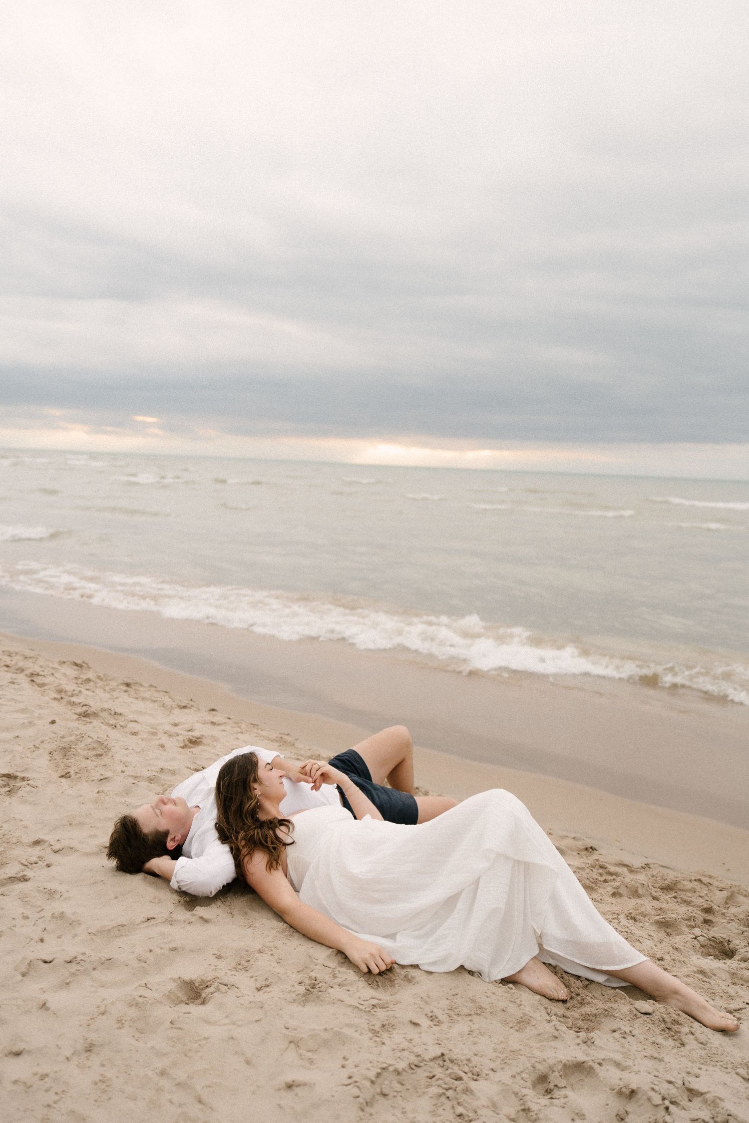 Romantic couple lying together on sandy beach near ocean waves under a dramatic cloudy sky at dusk.