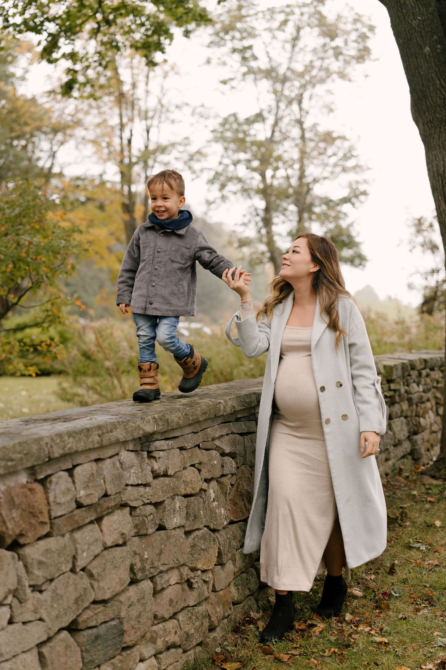 Pregnant woman in gray coat holds toddler son's hand as he walks on stone wall in autumn park.