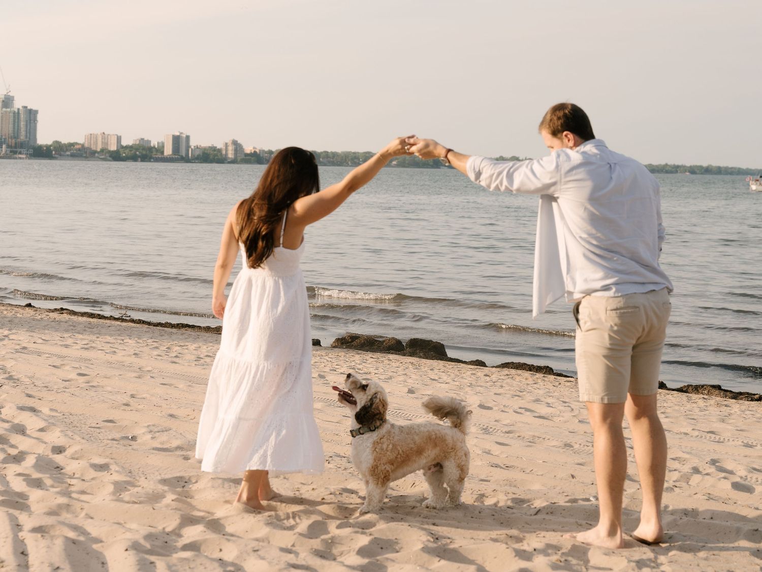 Couple dancing on beach with fluffy dog nearby and city skyline in background.