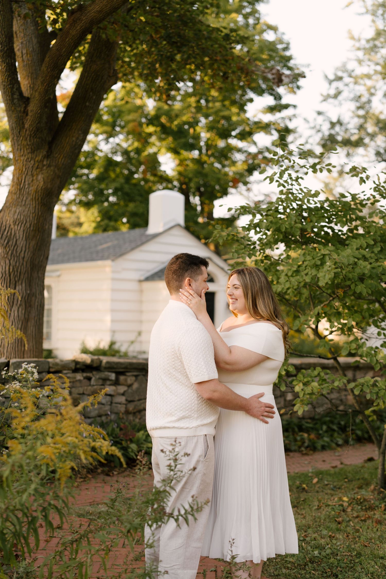 Couple in white outfits sharing a joyful embrace in a lush garden with a white cottage backdrop.