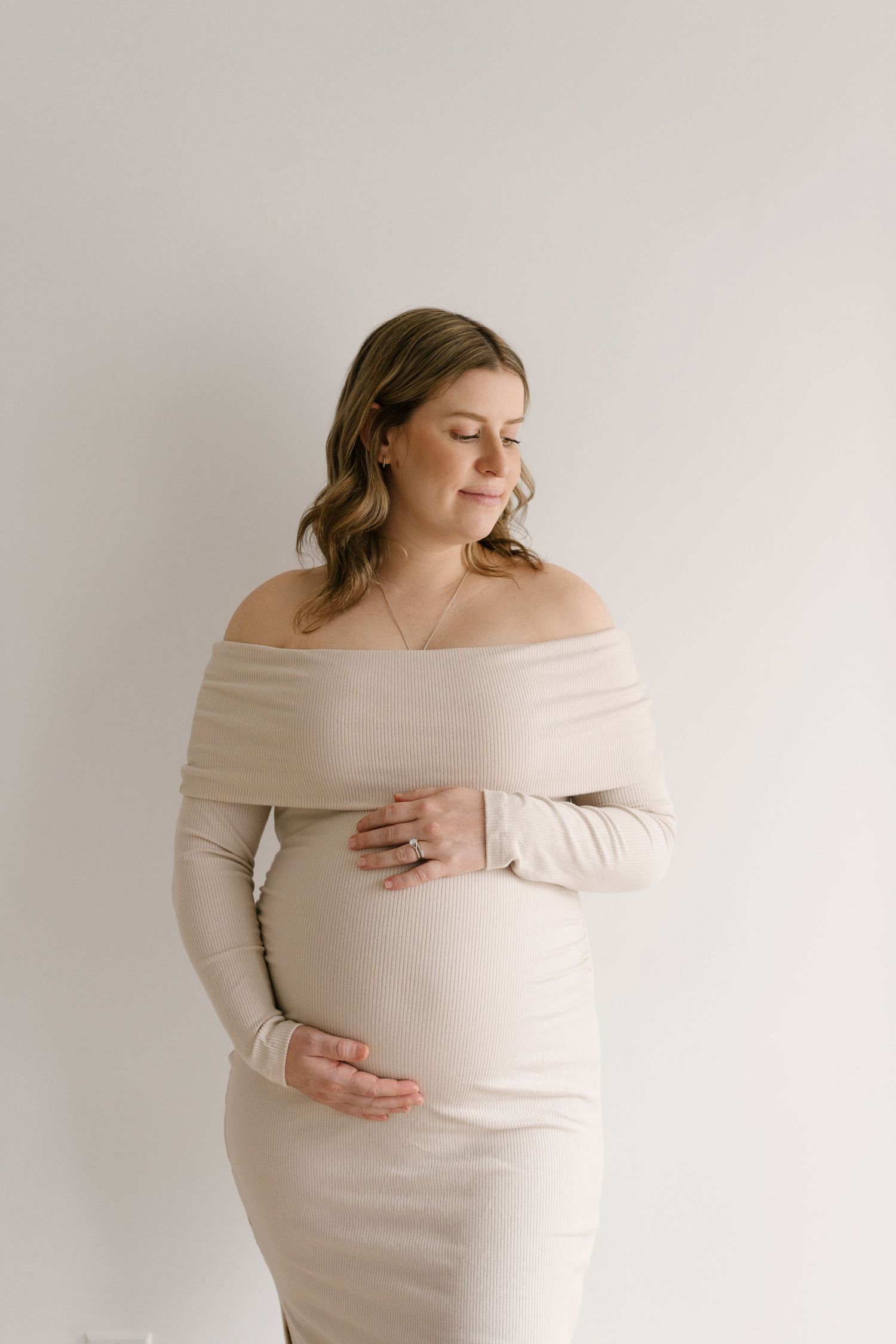 Pregnant woman in a beige off-shoulder ribbed dress gently cradling her baby bump against a white wall.