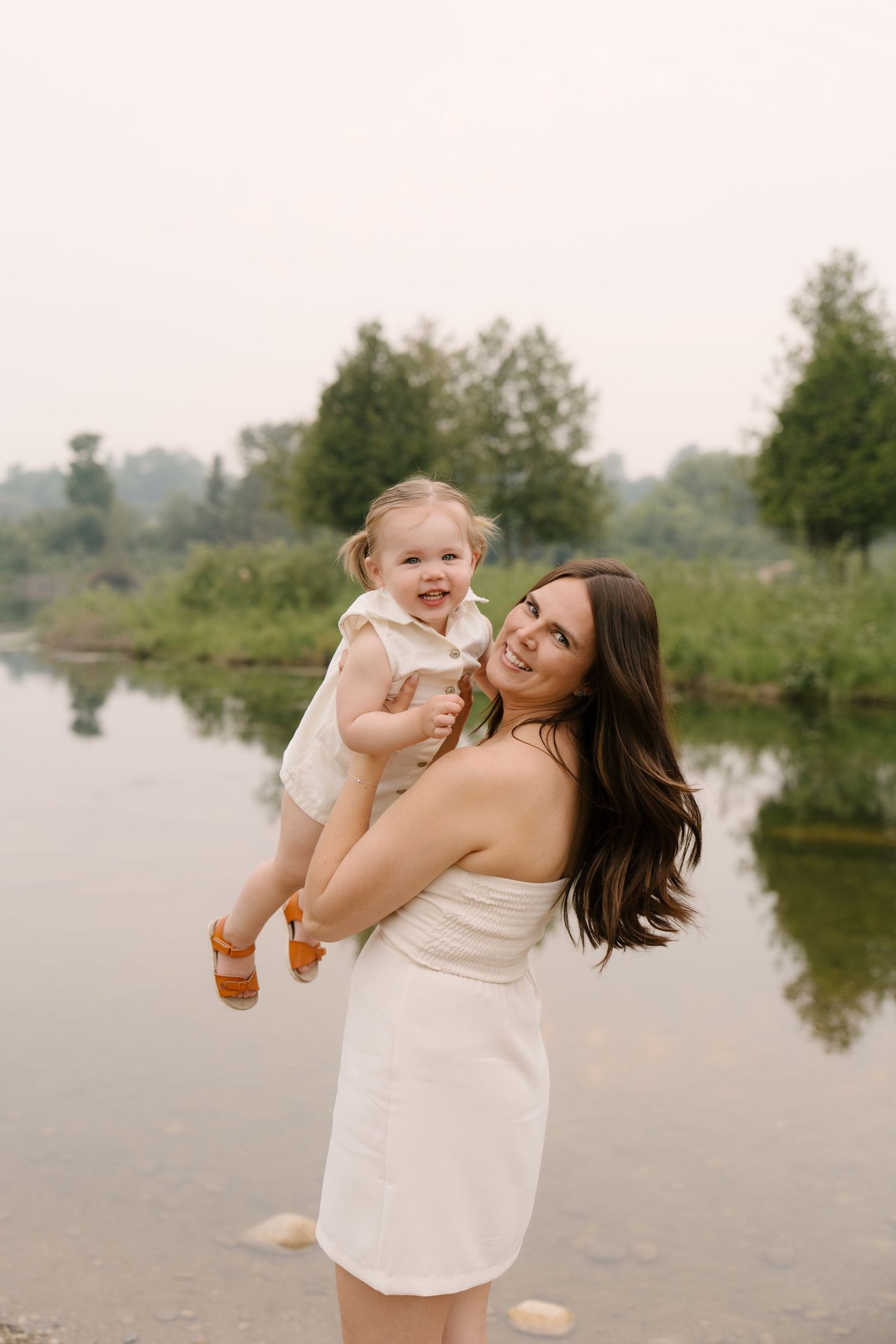 Happy mother in white dress lifting laughing toddler girl by a serene riverside.