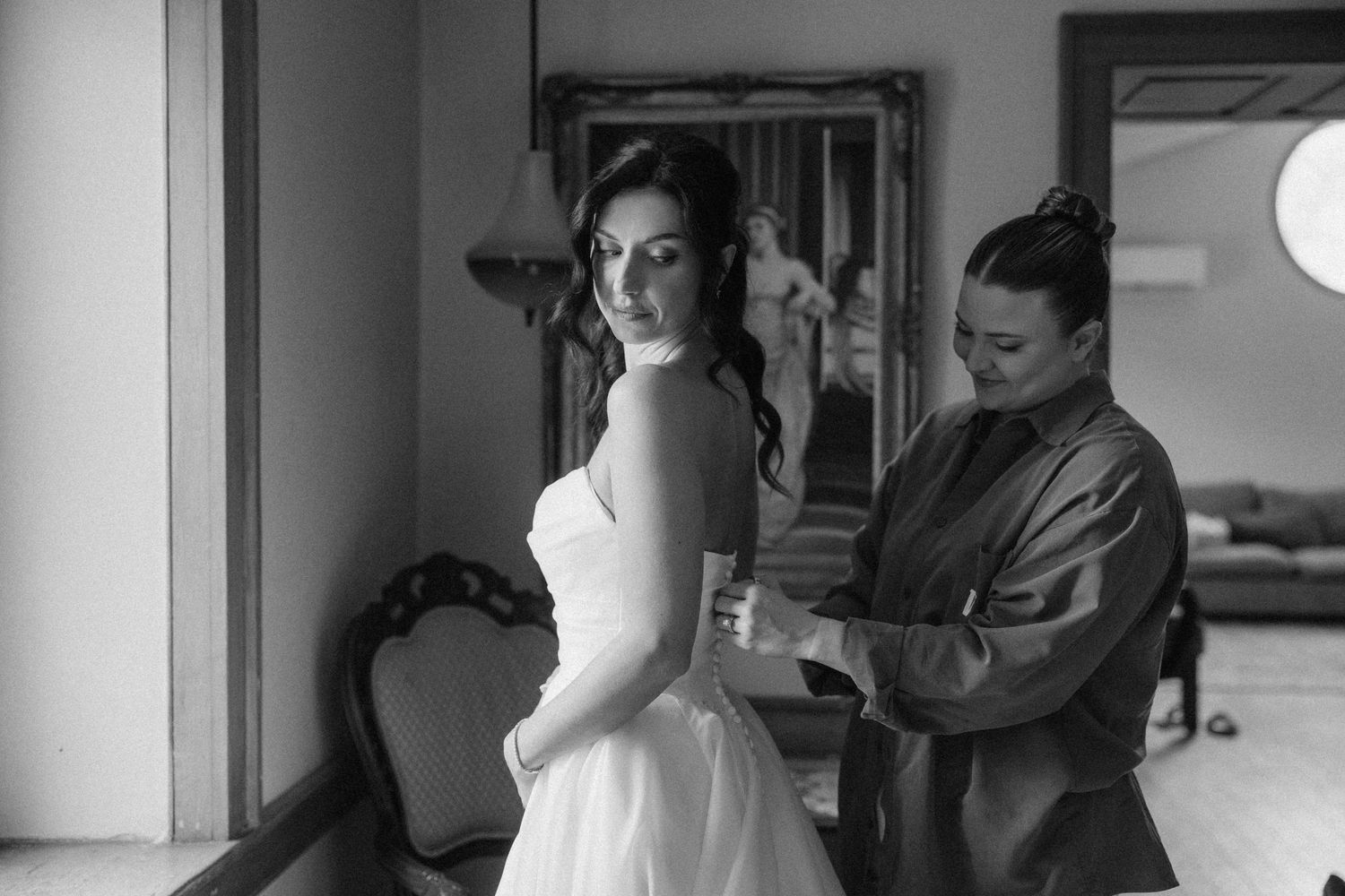 Black and white photo of bridesmaid helping bride fasten her strapless wedding gown indoors at Roseville Estates wedding venue.