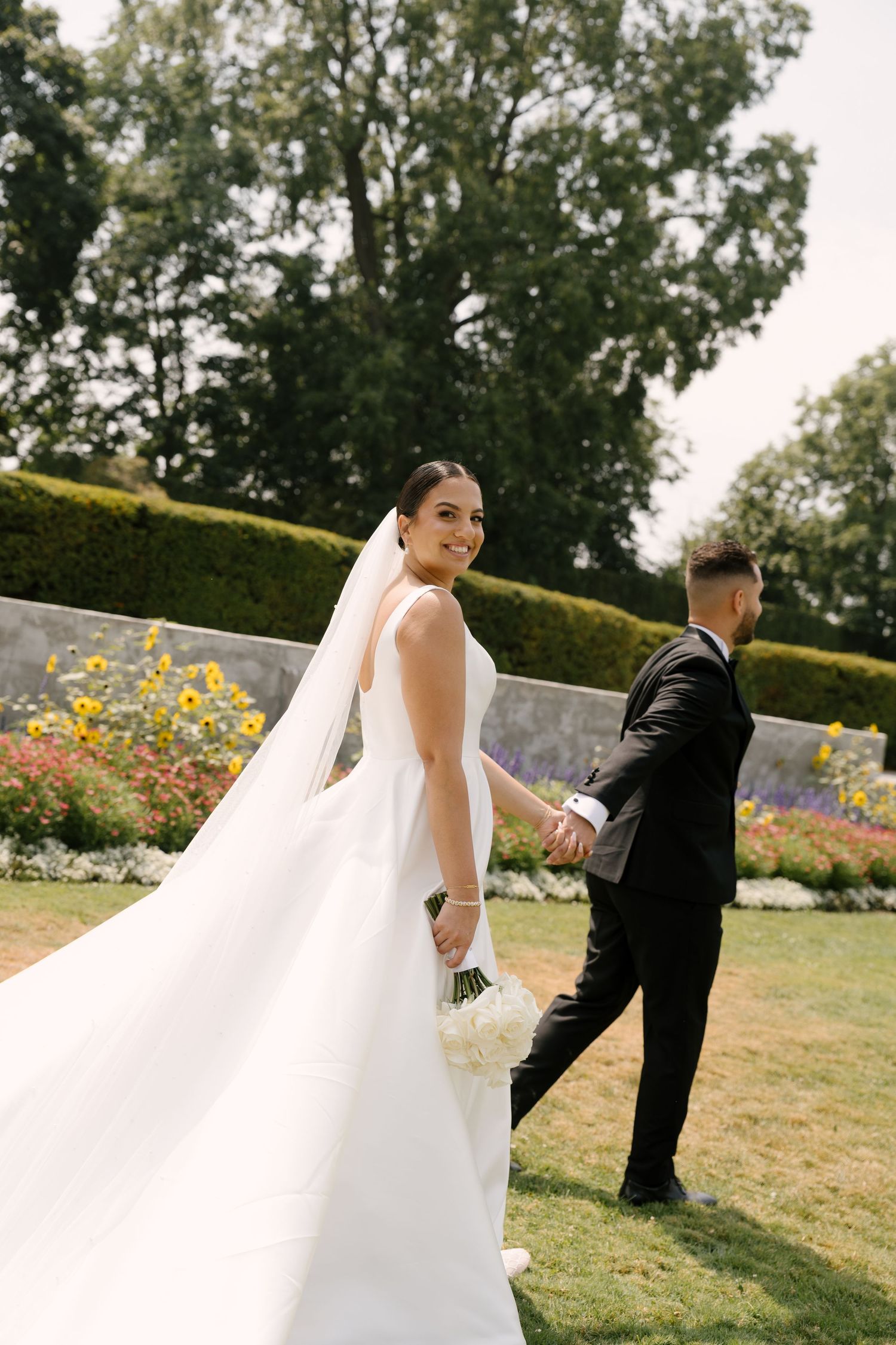 Bride in a flowing white gown with veil looks back smiling while leading groom through a formal garden setting at Parkwood Estates.