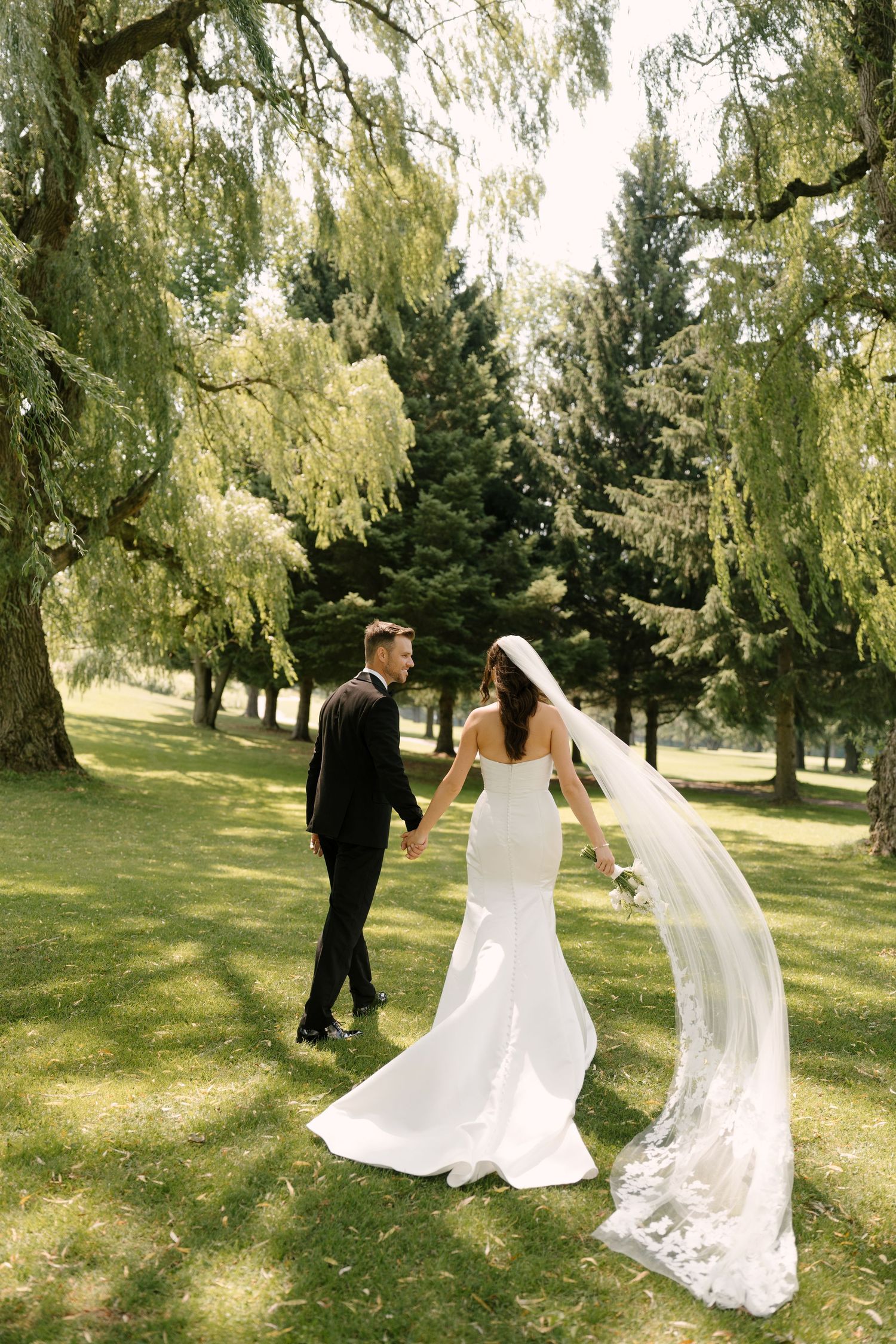 Bride and groom walk hand-in-hand through lush green park, bride wearing elegant mermaid gown with lace veil flowing.
