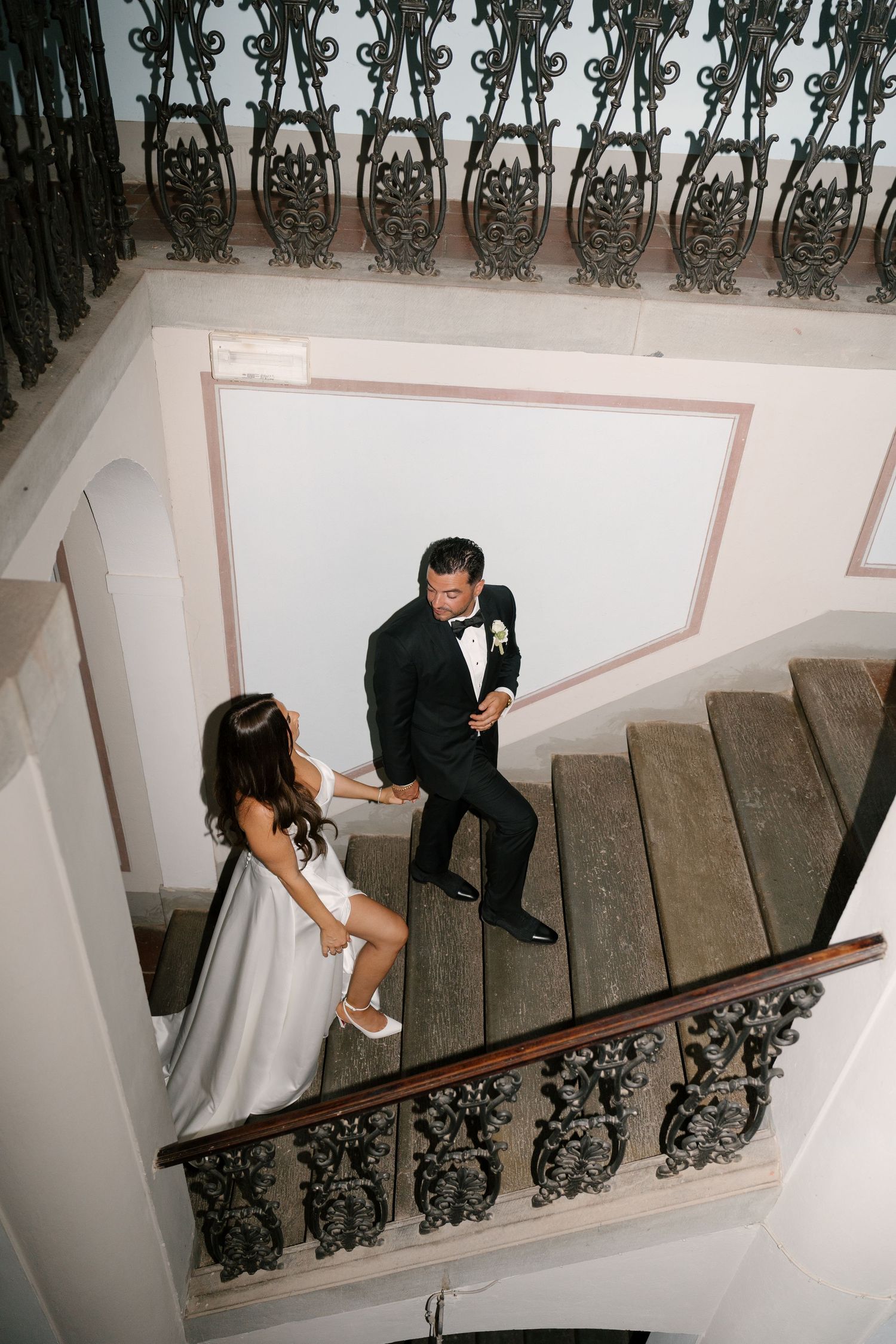 Bride and groom descend ornate wrought iron staircase inside elegant Italian villa wedding venue.