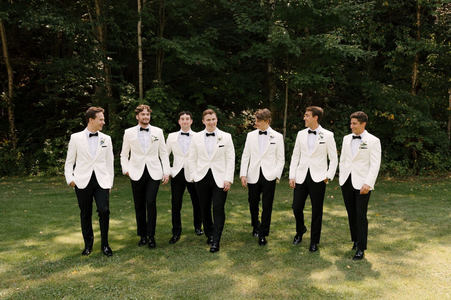 Seven groomsmen in white tuxedo jackets and black pants walk together on a green lawn with trees in the background.